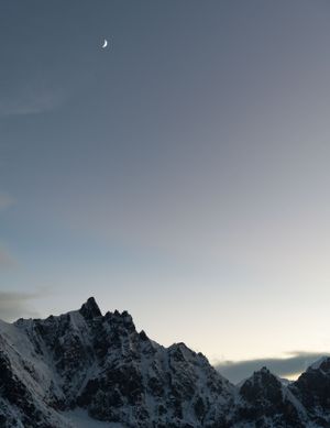 Crescent moon hanging over snow-covered mountain peaks at twilight in a winter landscape.