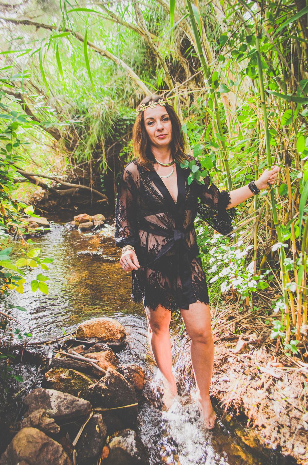 A model in a black lace coverup poses elegantly in a forest stream surrounded by bamboo and rocks.