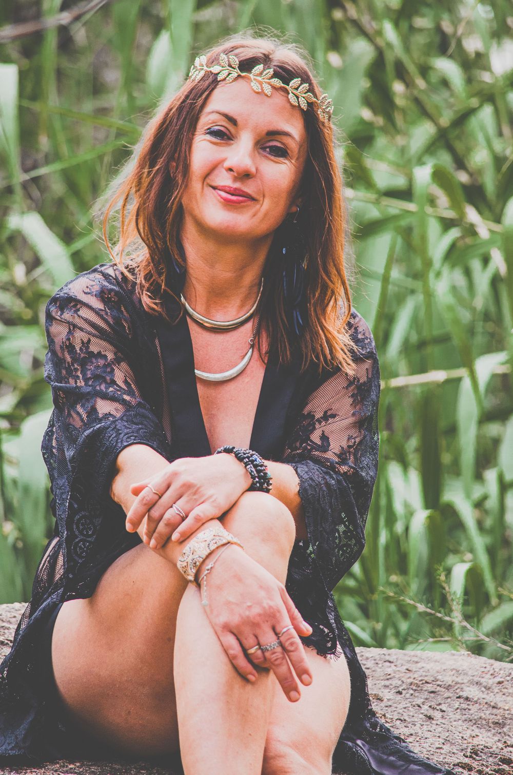 A relaxed outdoor portrait showing a gold crown, black lace dress, and jewelry against green foliage.