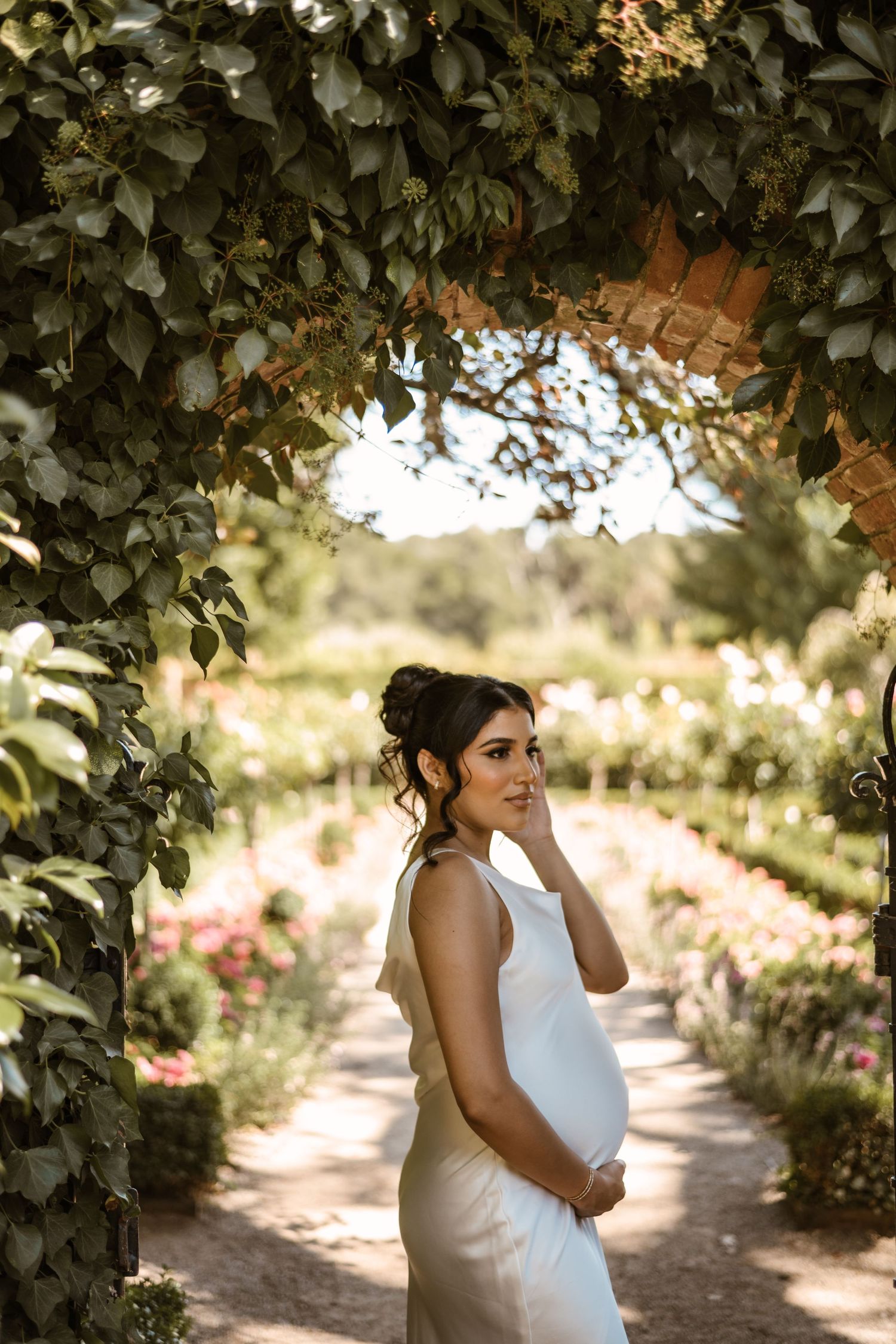 Bride in white dress stands thoughtfully in garden path surrounded by lush greenery.
