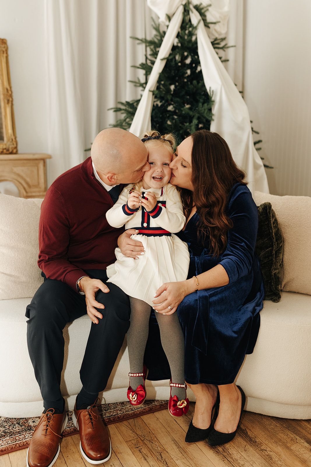 Family shares holiday moment together sitting on white couch with Christmas tree in background.