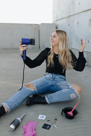 Vlogger sits on ground with hair dryer and styling tools while filming a beauty tutorial in casual attire.