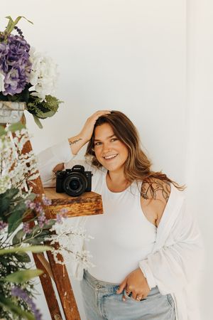 Photographer poses casually near floral arrangement with camera and natural decor on wooden ladder.