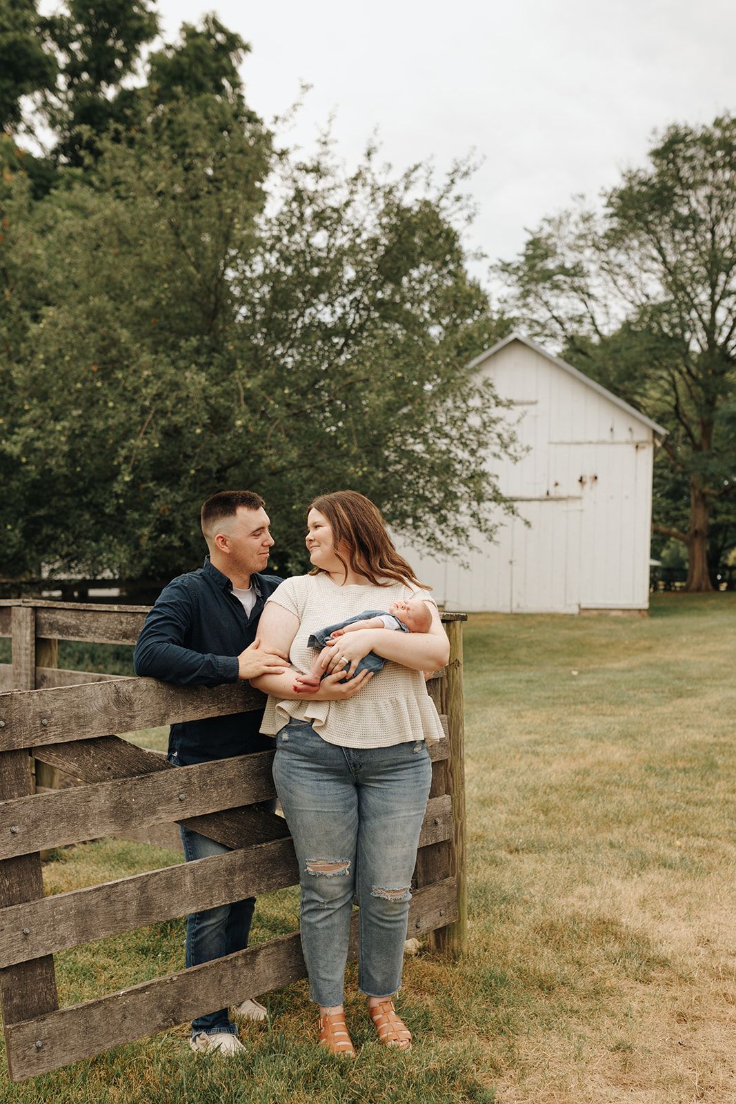 Couple shares a tender moment with their newborn while leaning against a rustic wooden fence with a white barn in the background.