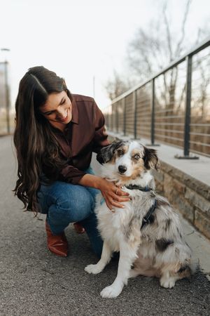 A person in a brown shirt and jeans kneels beside their Australian Shepherd mix dog on a paved walkway.