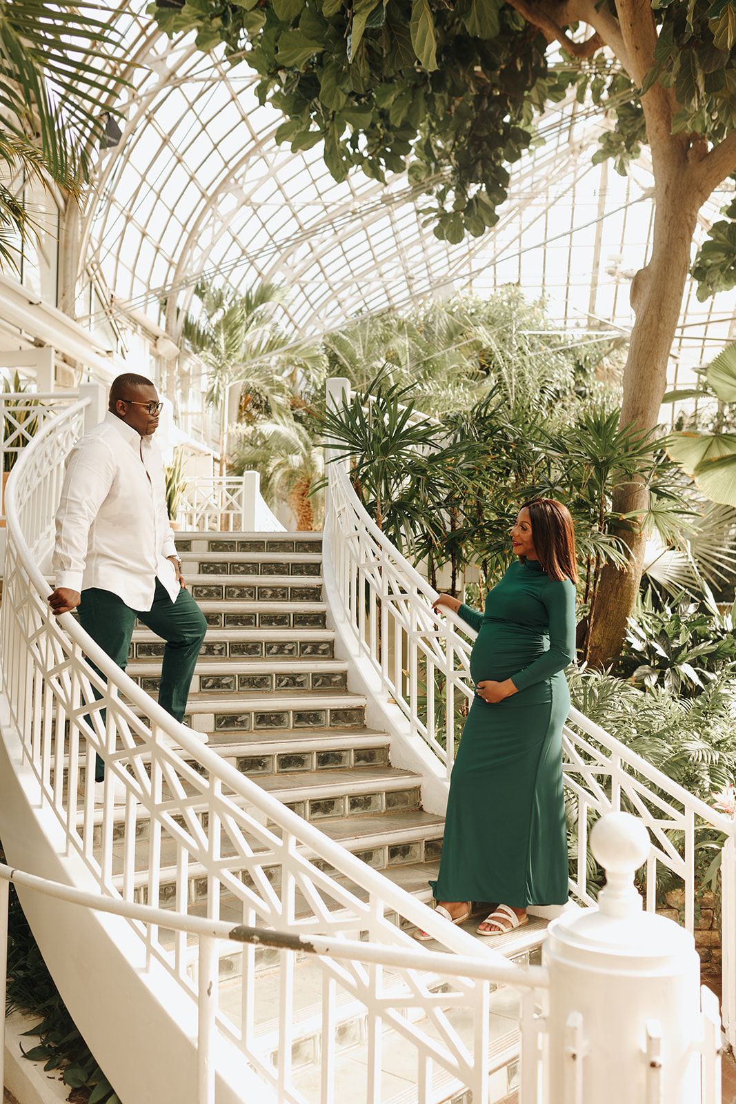 A couple poses on elegant white stairs inside a greenhouse-style conservatory filled with tropical plants and natural light.