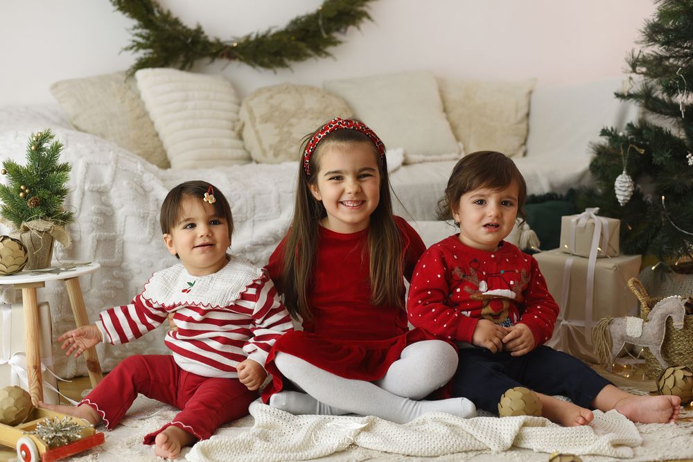 Three children in festive red and white outfits sit together among Christmas decorations and wrapped presents.