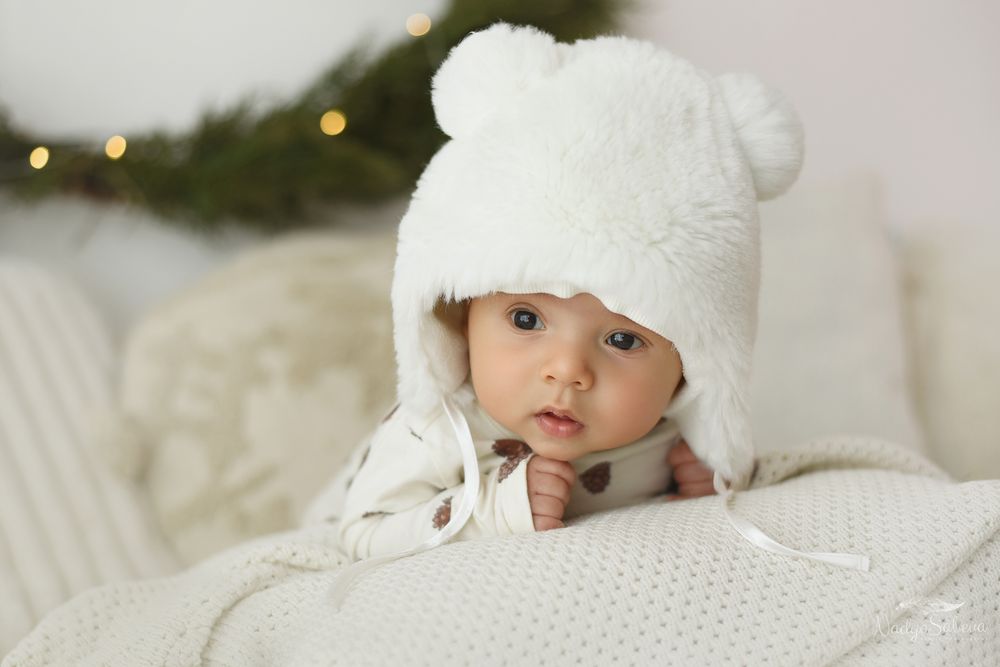 A baby wearing a white winter hat lies on soft white bedding while looking at the camera.