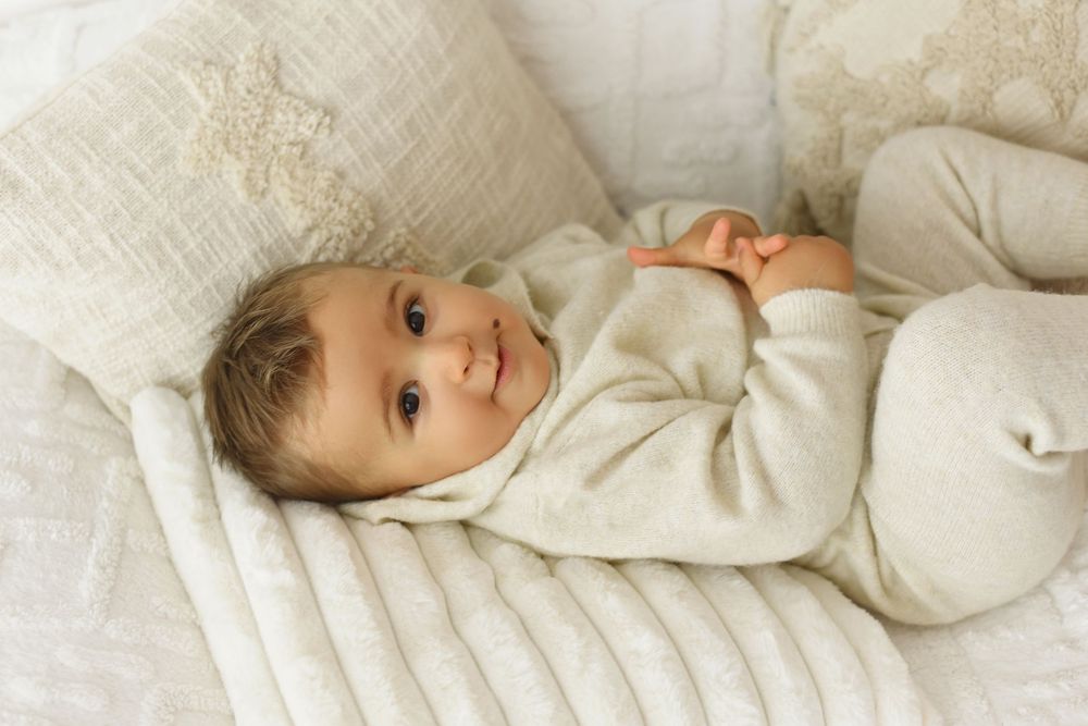A young child in cream-colored clothing lies on white bedding with a bright smile.
