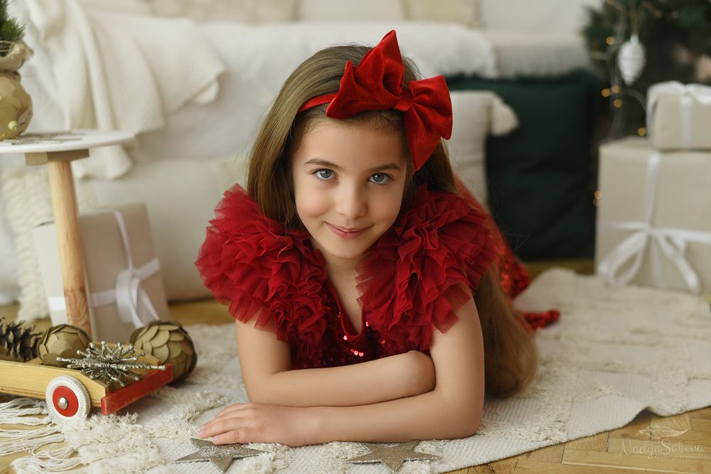 A young child in a festive red outfit with a matching bow lies on a white blanket during a holiday photo session.