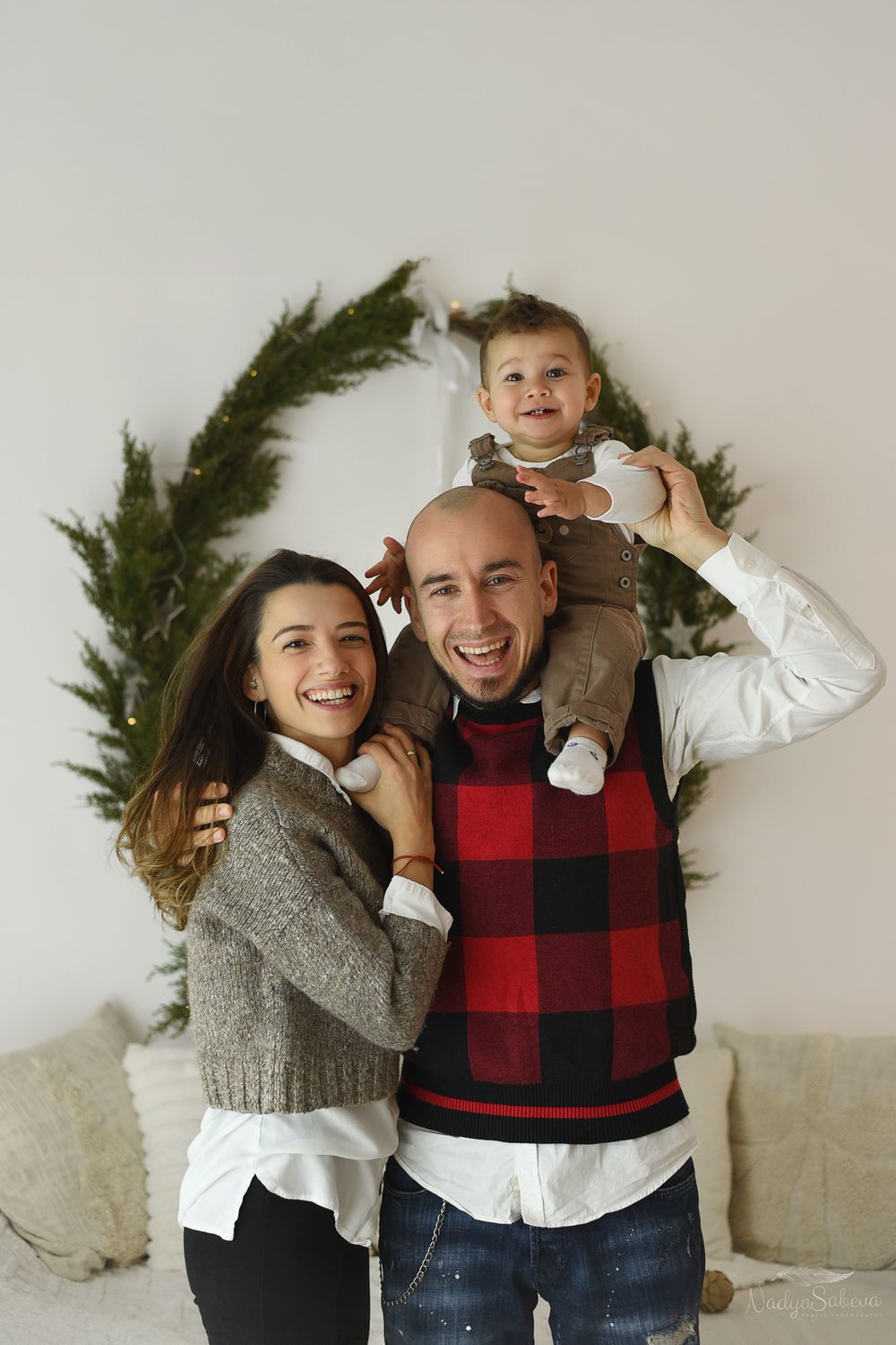 Happy family poses together in front of evergreen wreath during holiday photoshoot wearing cozy winter sweaters.