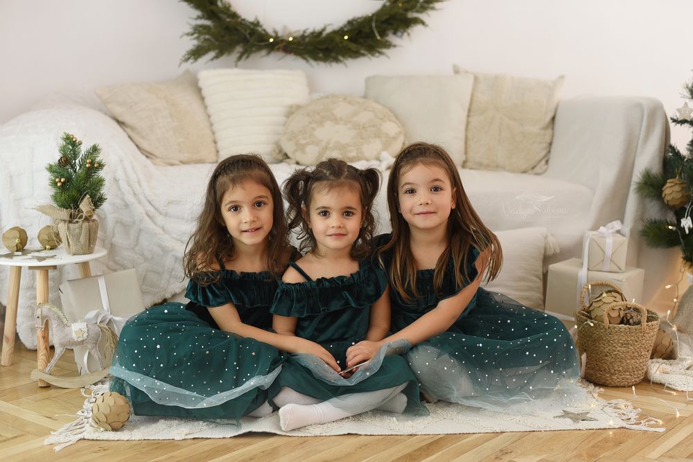 Three children in matching dark dresses with sparkly tulle skirts sit together on floor with holiday decor in background.