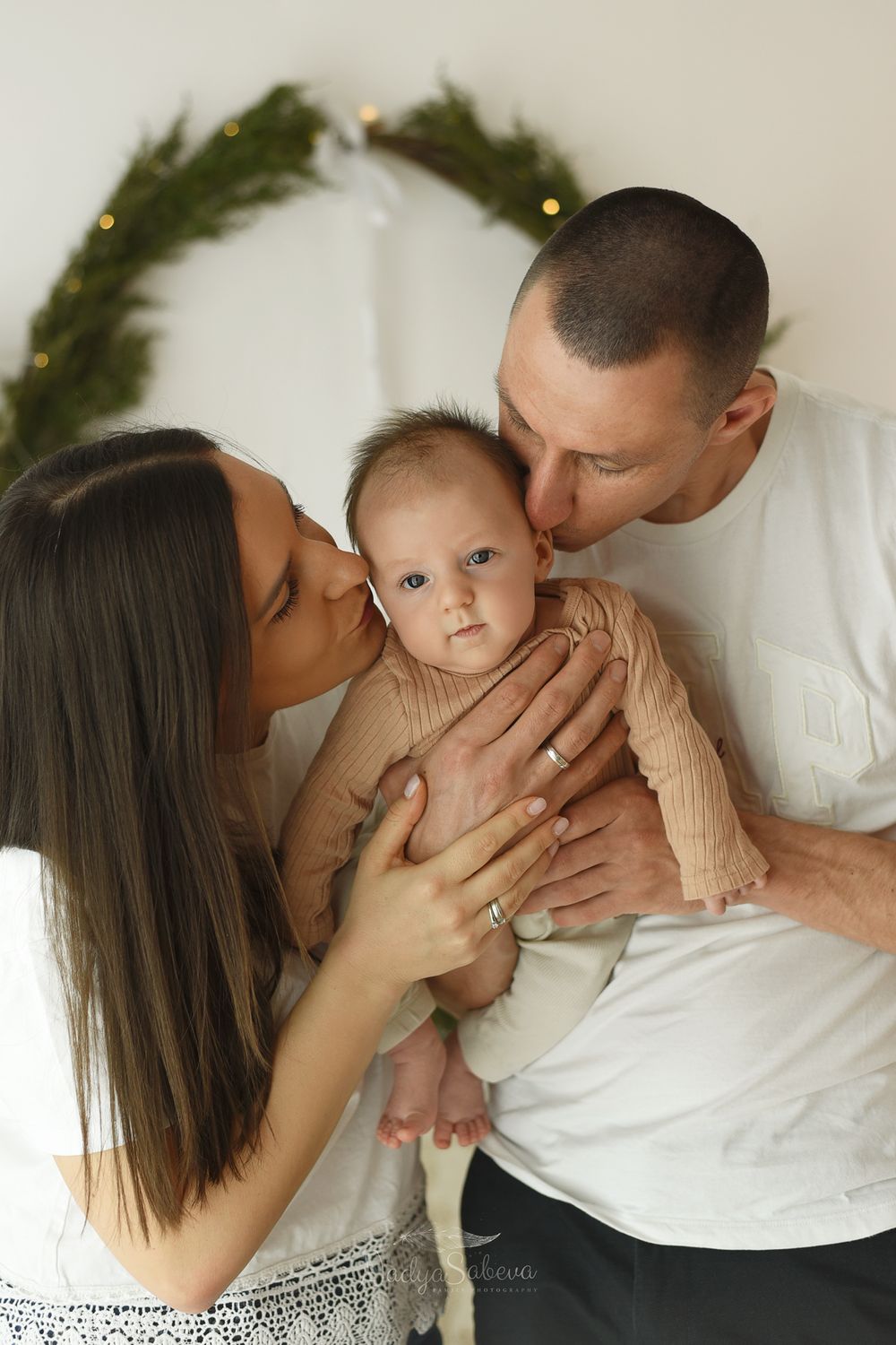 A loving family moment captured during a holiday photoshoot with a baby and parents against a white background with evergreen decor.