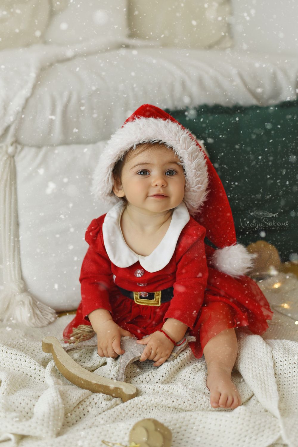 A baby wearing a red Santa outfit with white fur trim lies on a white blanket during a festive holiday photoshoot.