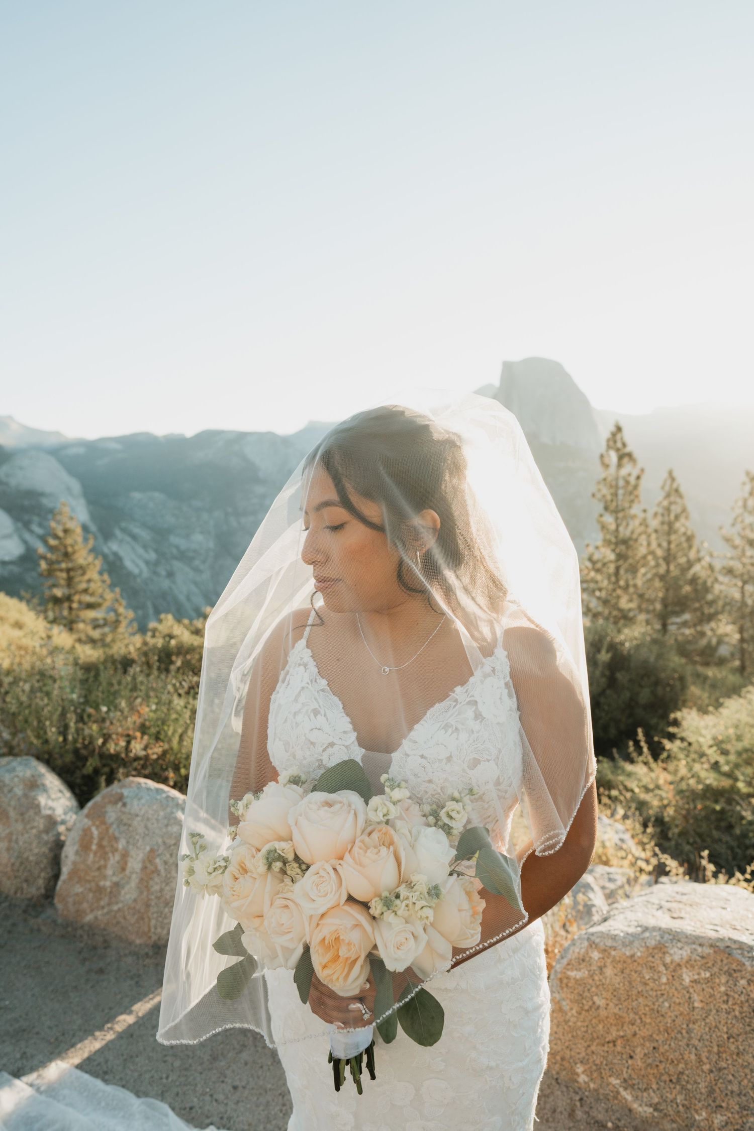 A bride holding a bouquet poses at a scenic mountain overlook during golden hour.