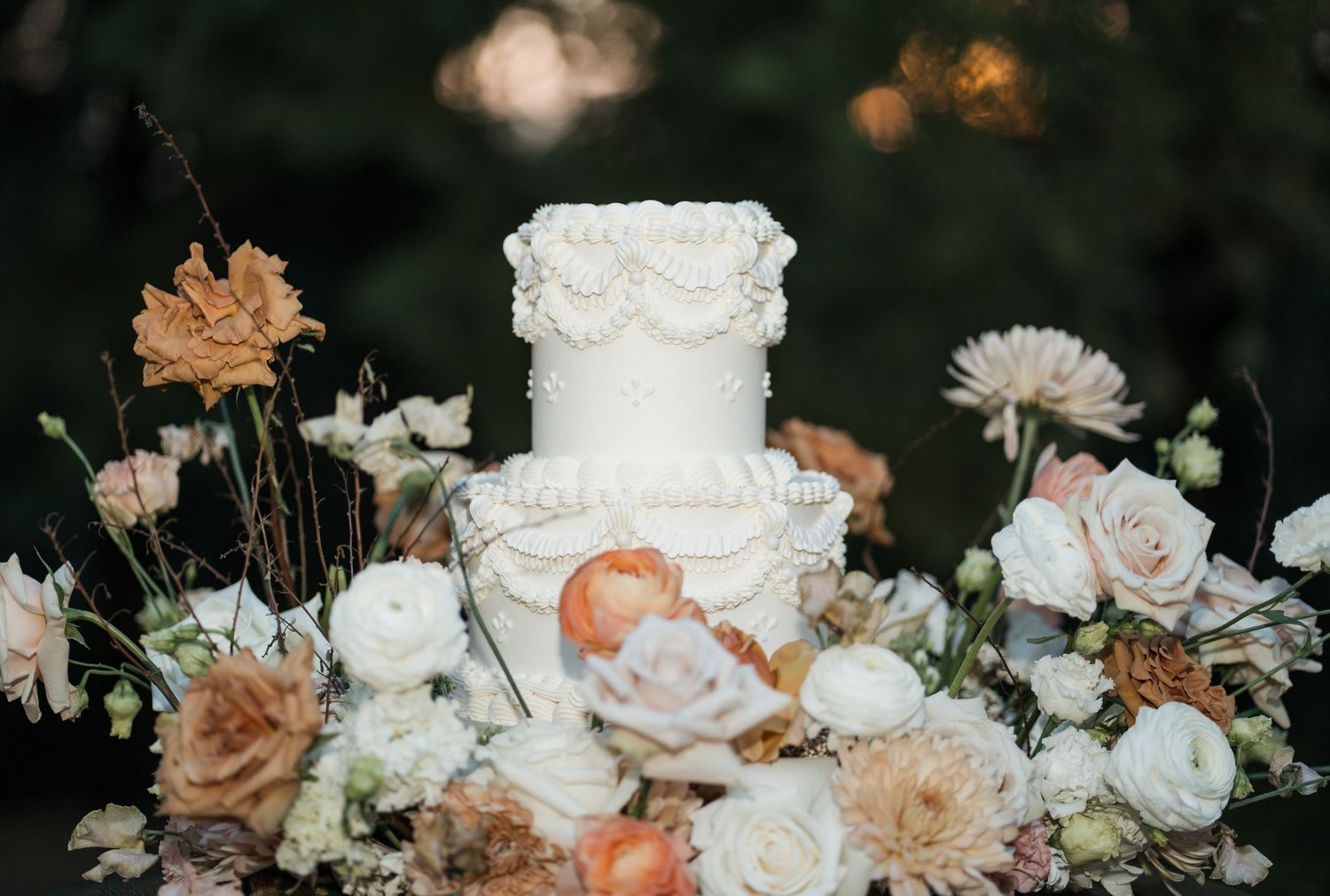 An elegant white wedding cake decorated with roses and eucalyptus with a golden topper.