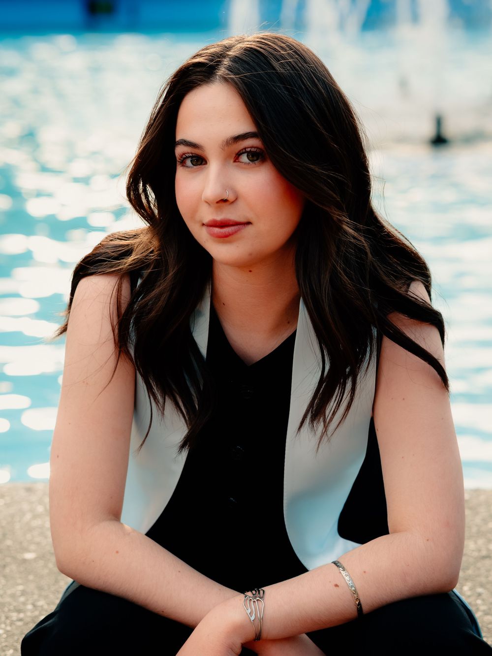 Graduate in black and white attire sits by a blue fountain during a celebration photoshoot.