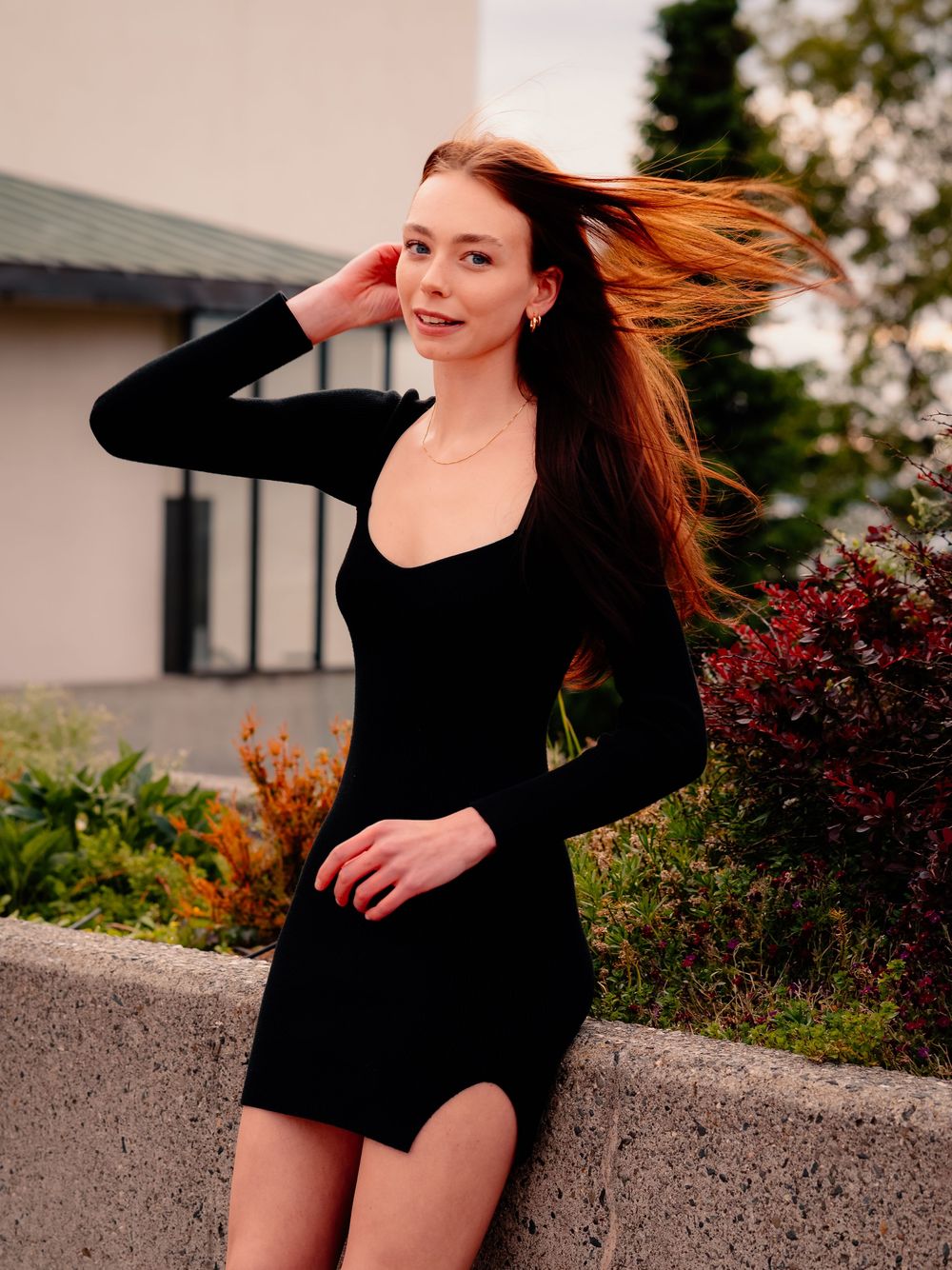 A graduate in a black dress poses with windswept hair against autumn foliage.