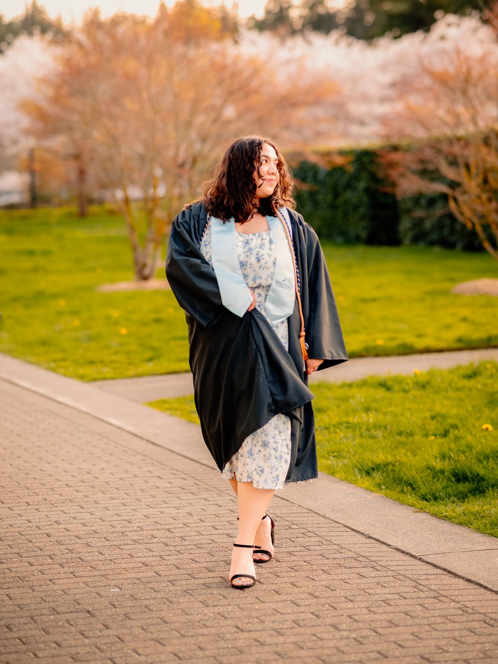 Walking shot of graduate in flowing gown and floral dress on campus pathway.