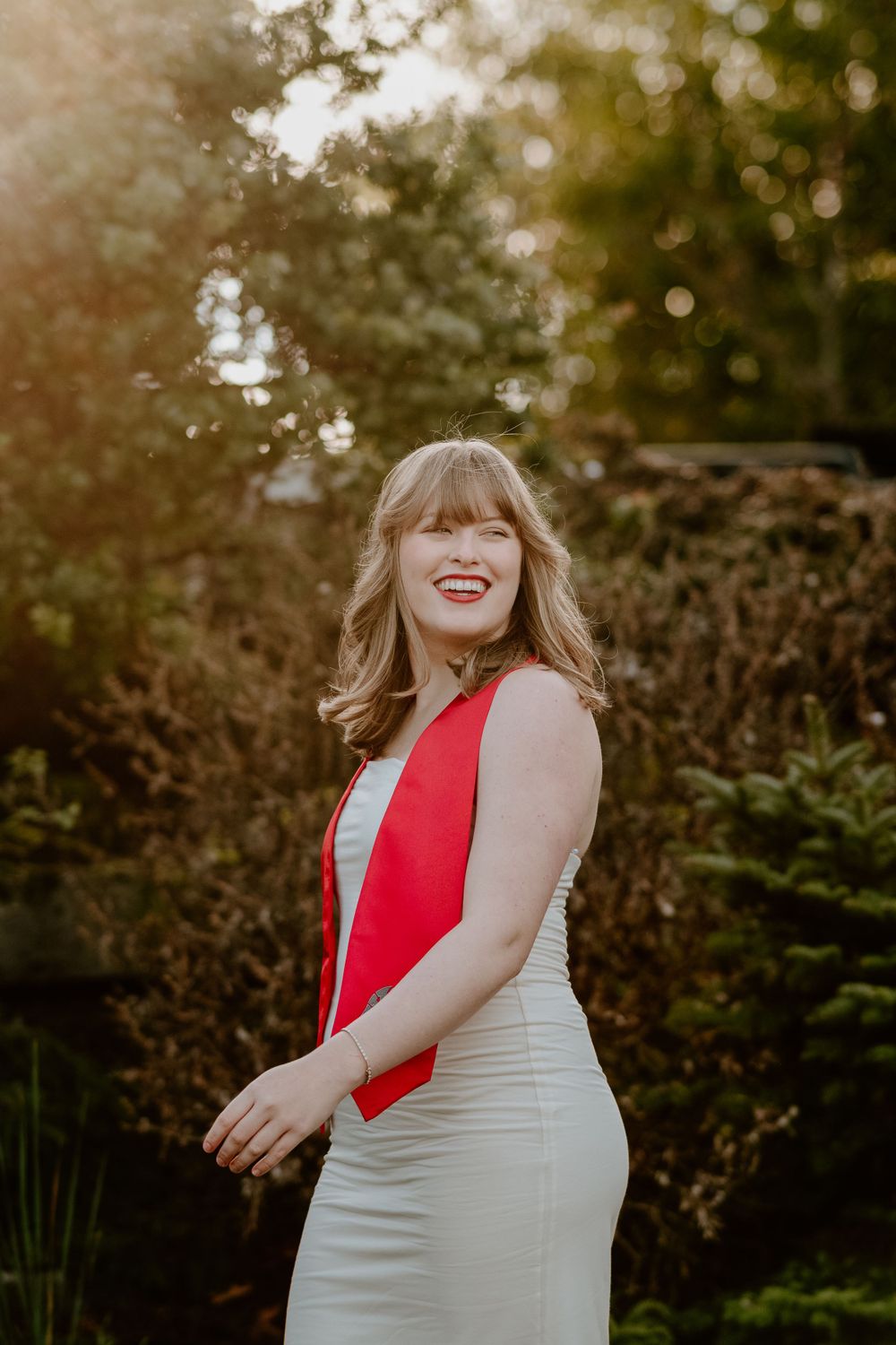A person in a white dress with red accents smiles joyfully during an outdoor photoshoot at sunset.