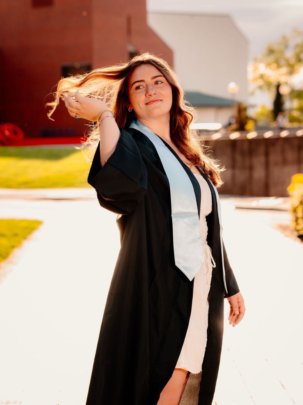 Graduate in cap and gown poses triumphantly in sunlit outdoor setting.
