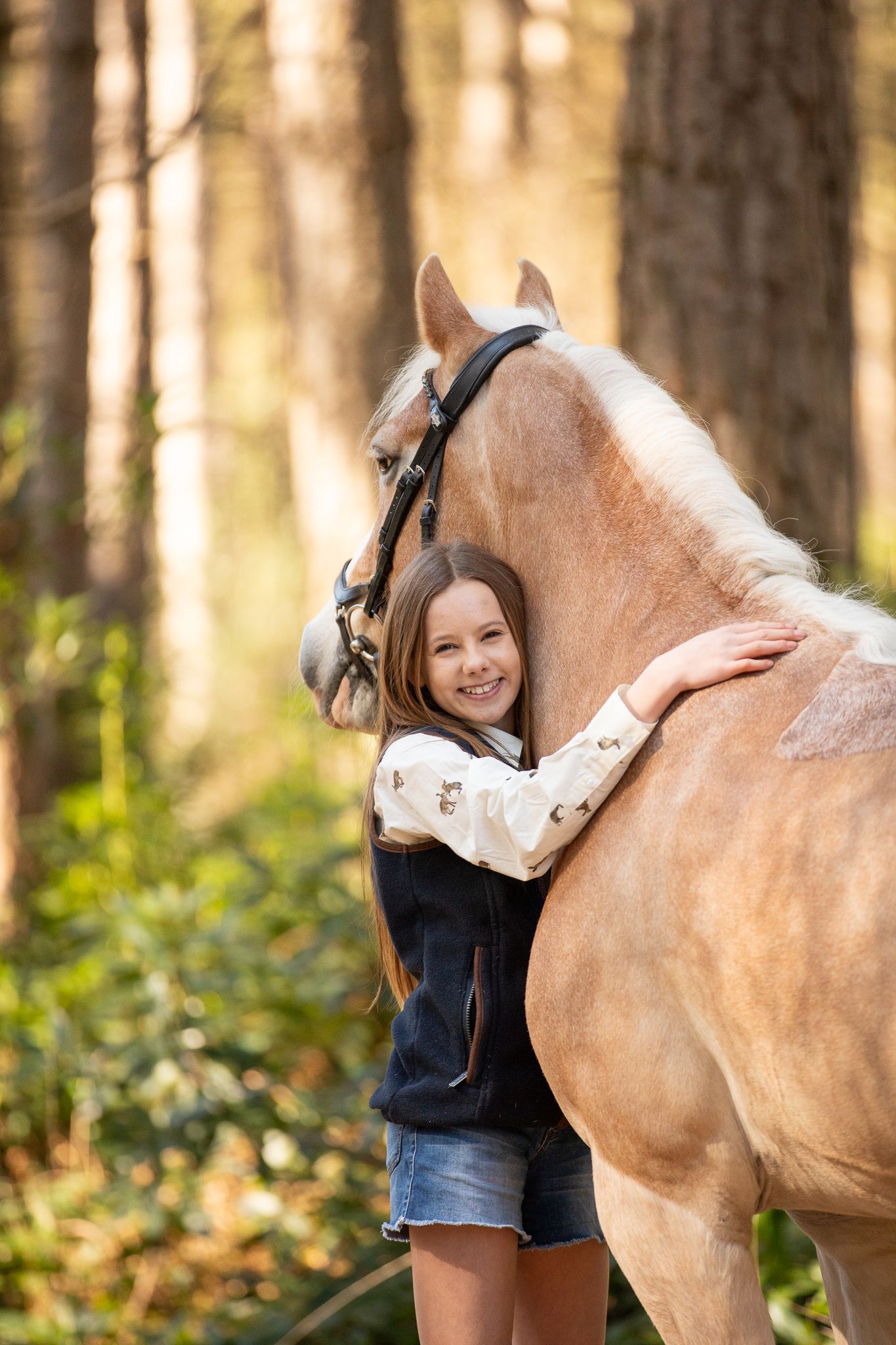 Young girl in denim overalls embraces a palomino horse in a sun-dappled forest setting.