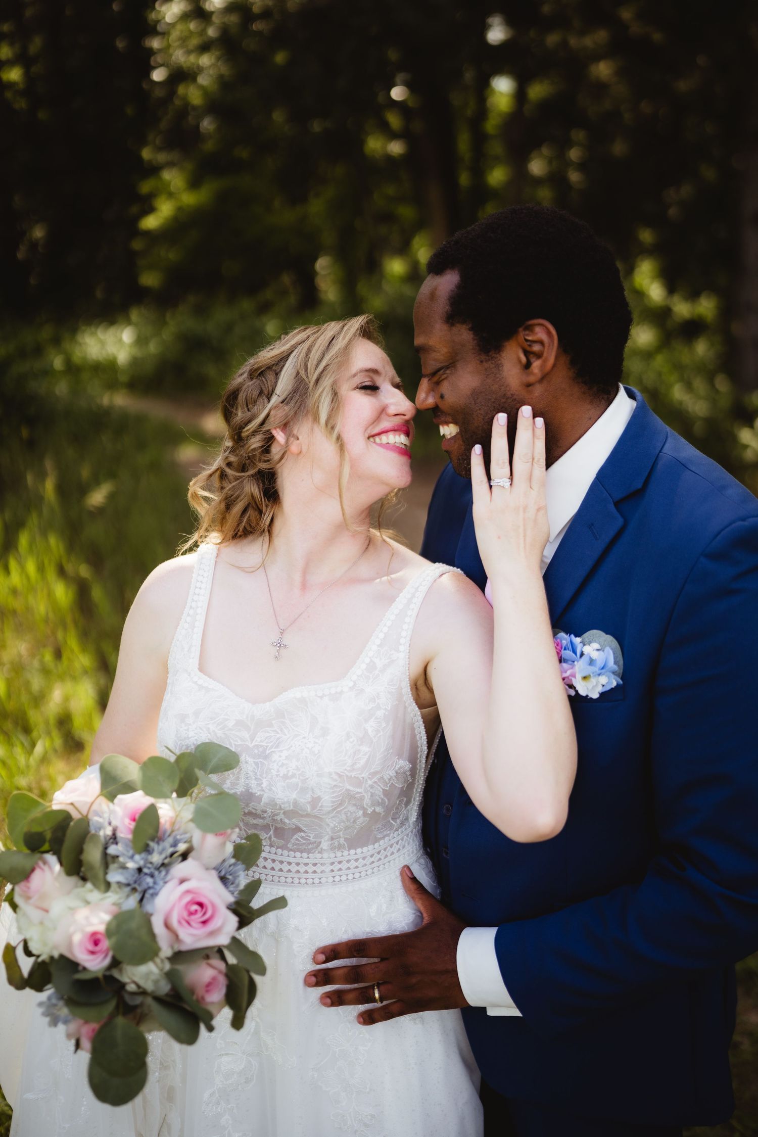 Happy newlywed couple embraces outdoors while bride holds pink and white floral bouquet in summer garden setting.