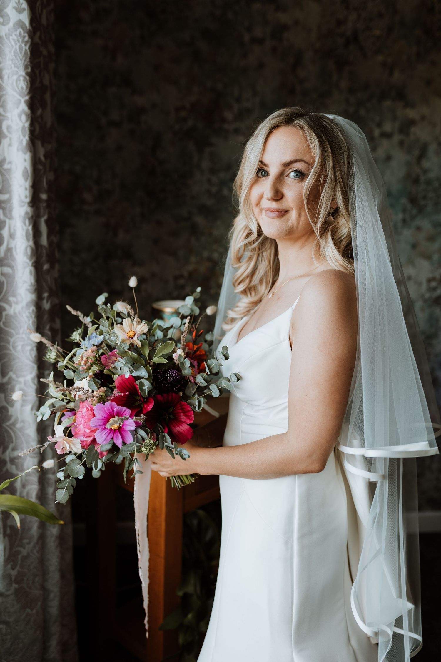 A bride in a sleek white wedding dress holds a vibrant bouquet with red and pink flowers against a dark textured background.