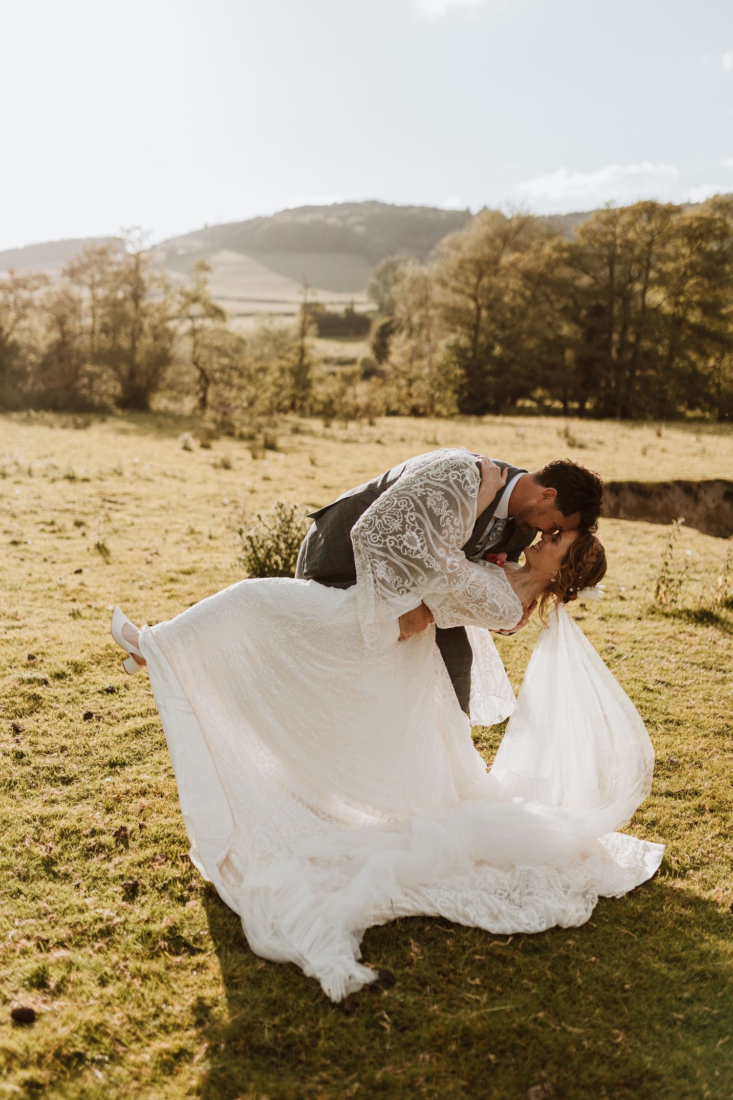 A romantic countryside wedding scene shows a couple embracing on a grassy hillside at sunset.