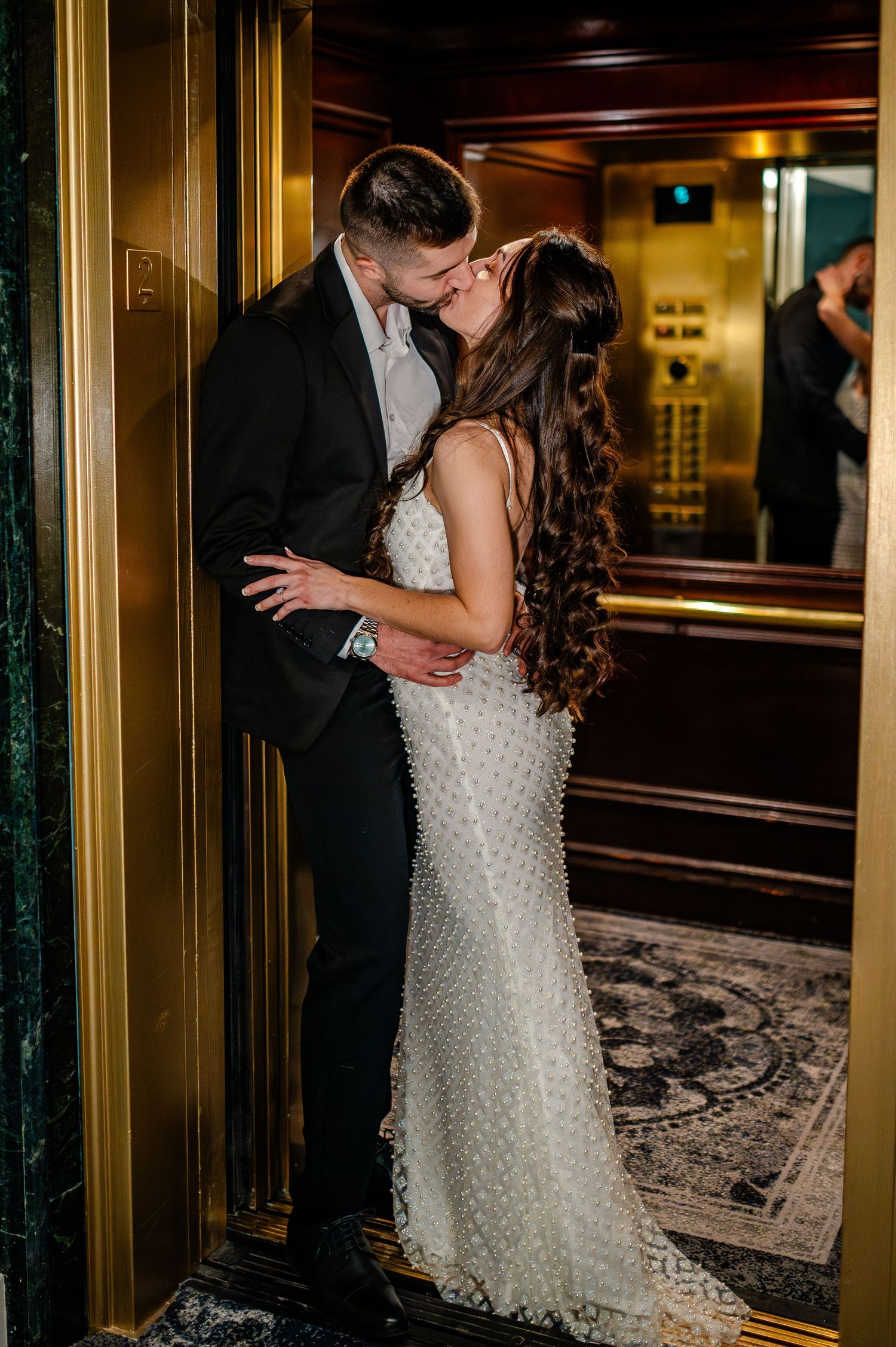 A bride in a white beaded gown and dark-suited partner embrace in a dimly lit elegant hallway.