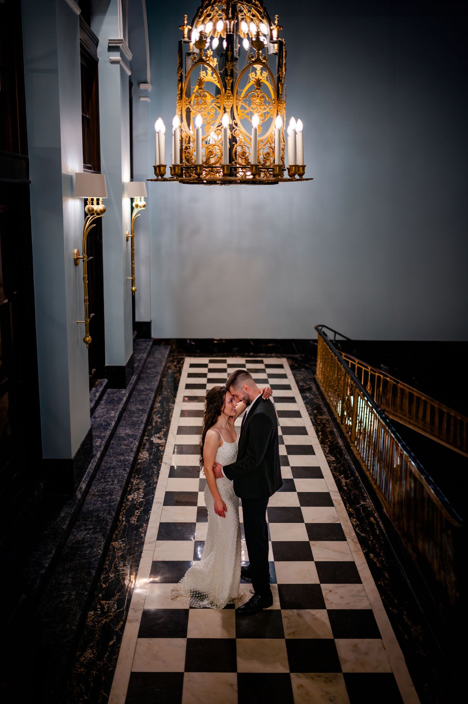 A romantic scene under an ornate gold chandelier on a black and white checkered floor in an elegant interior.