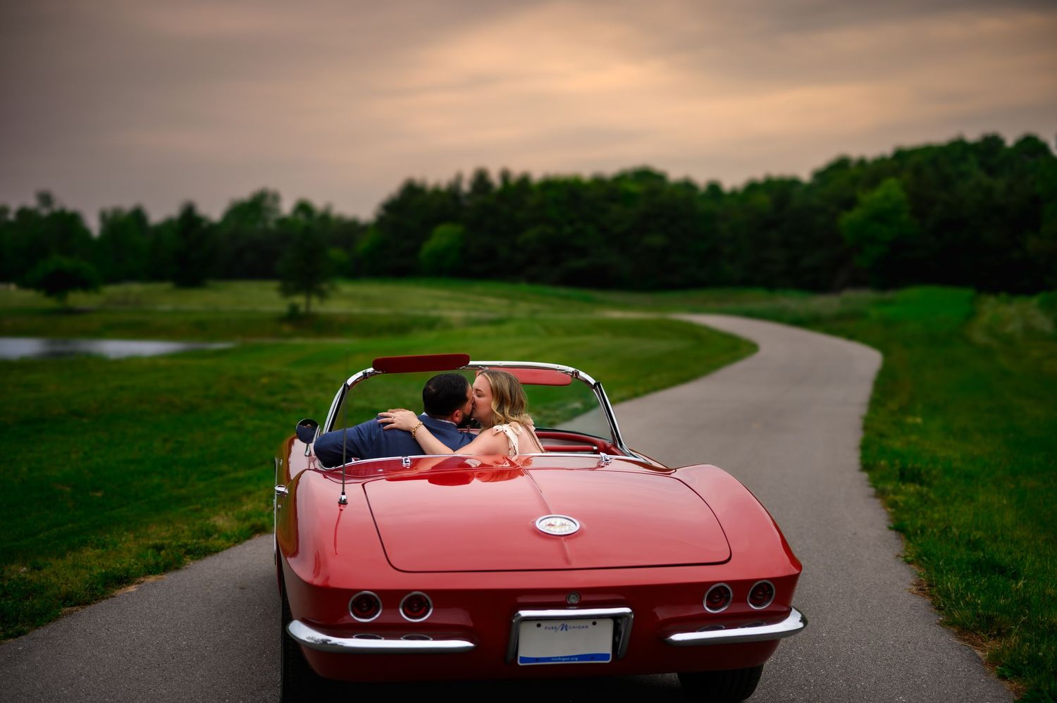 A vintage red Chevrolet Corvette convertible drives along a winding country road surrounded by green landscape.