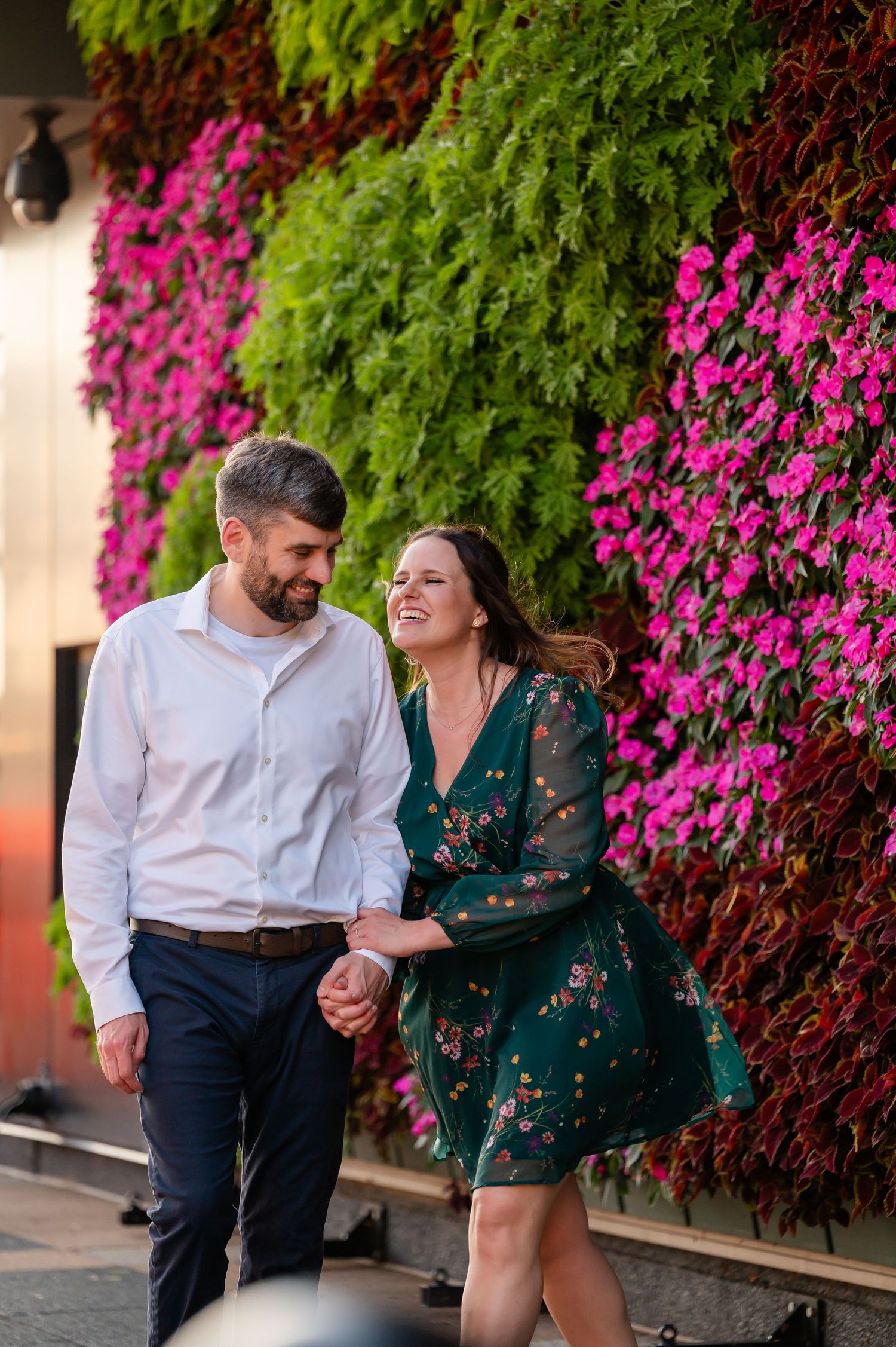A couple shares a romantic moment together against a wall covered in vibrant pink flowers and green foliage.