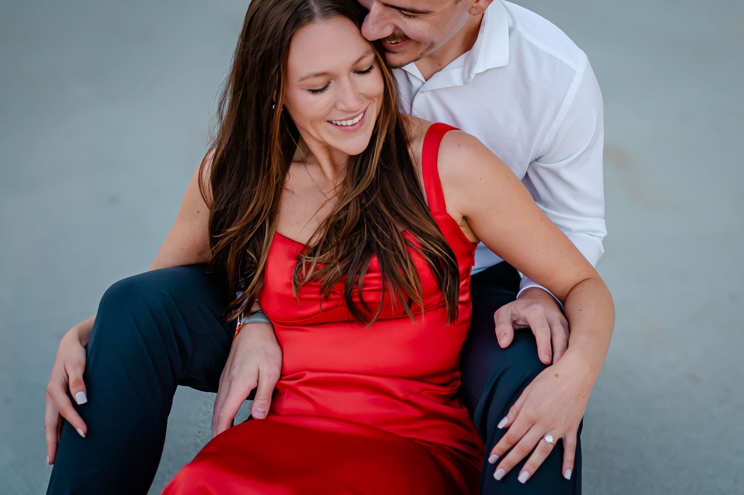 A romantic couple in red dress and white shirt embrace and laugh together during a portrait photo session.