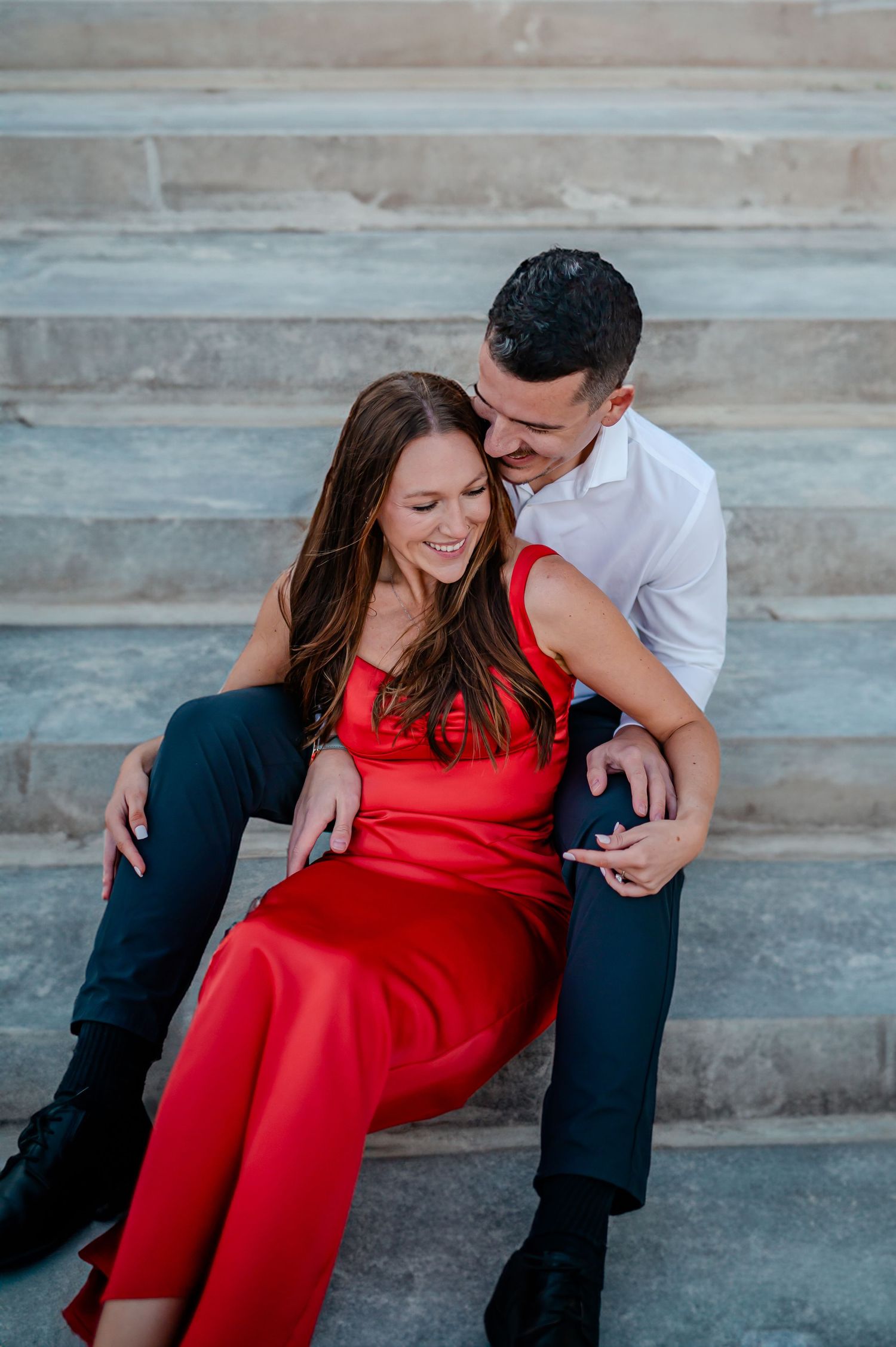 Romantic couple in love sitting on stone stairs, woman wearing a red dress and man in dark suit embracing tenderly.