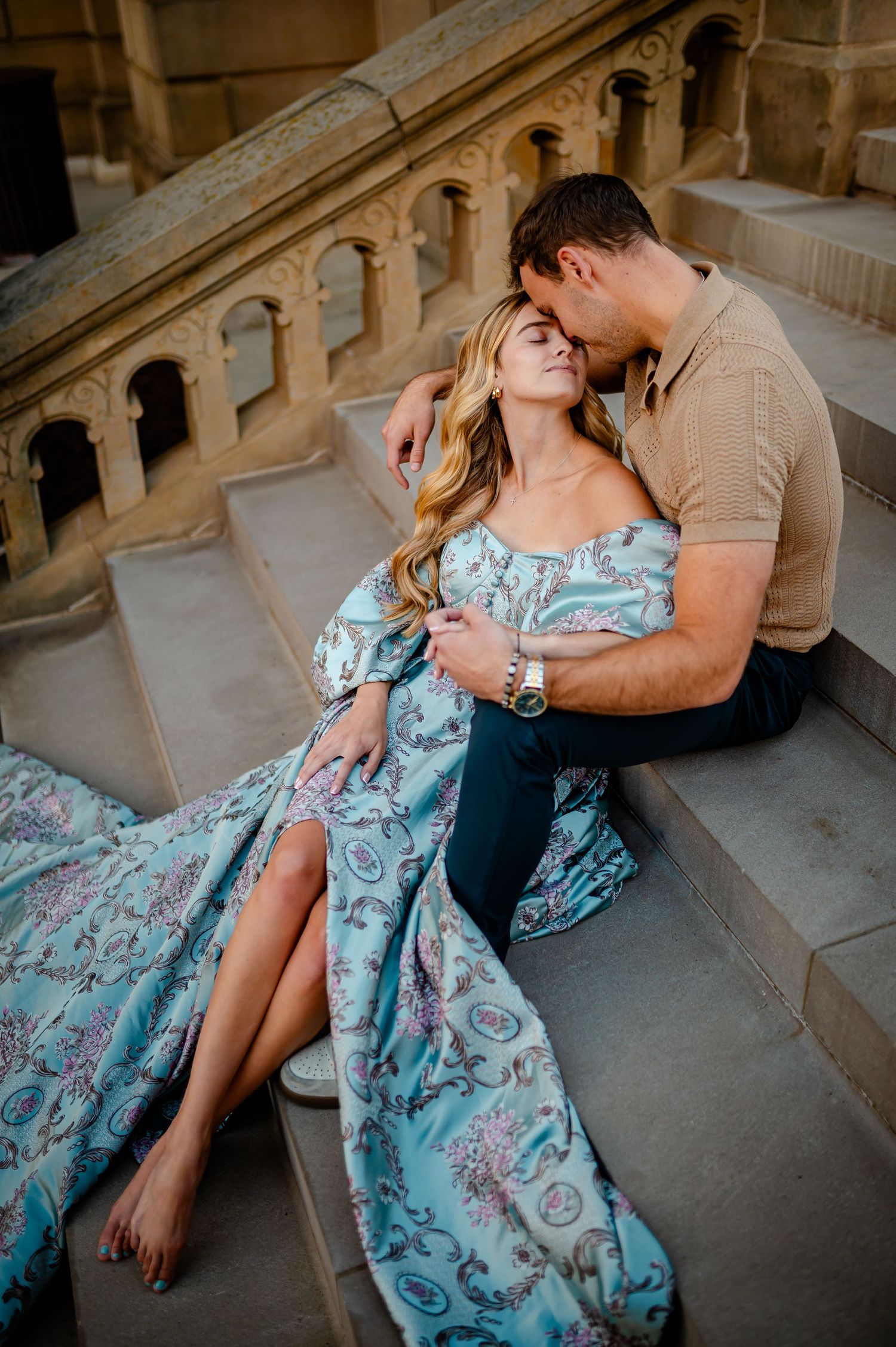 A couple shares an intimate moment together on stone steps as she wears a flowing blue floral dress.