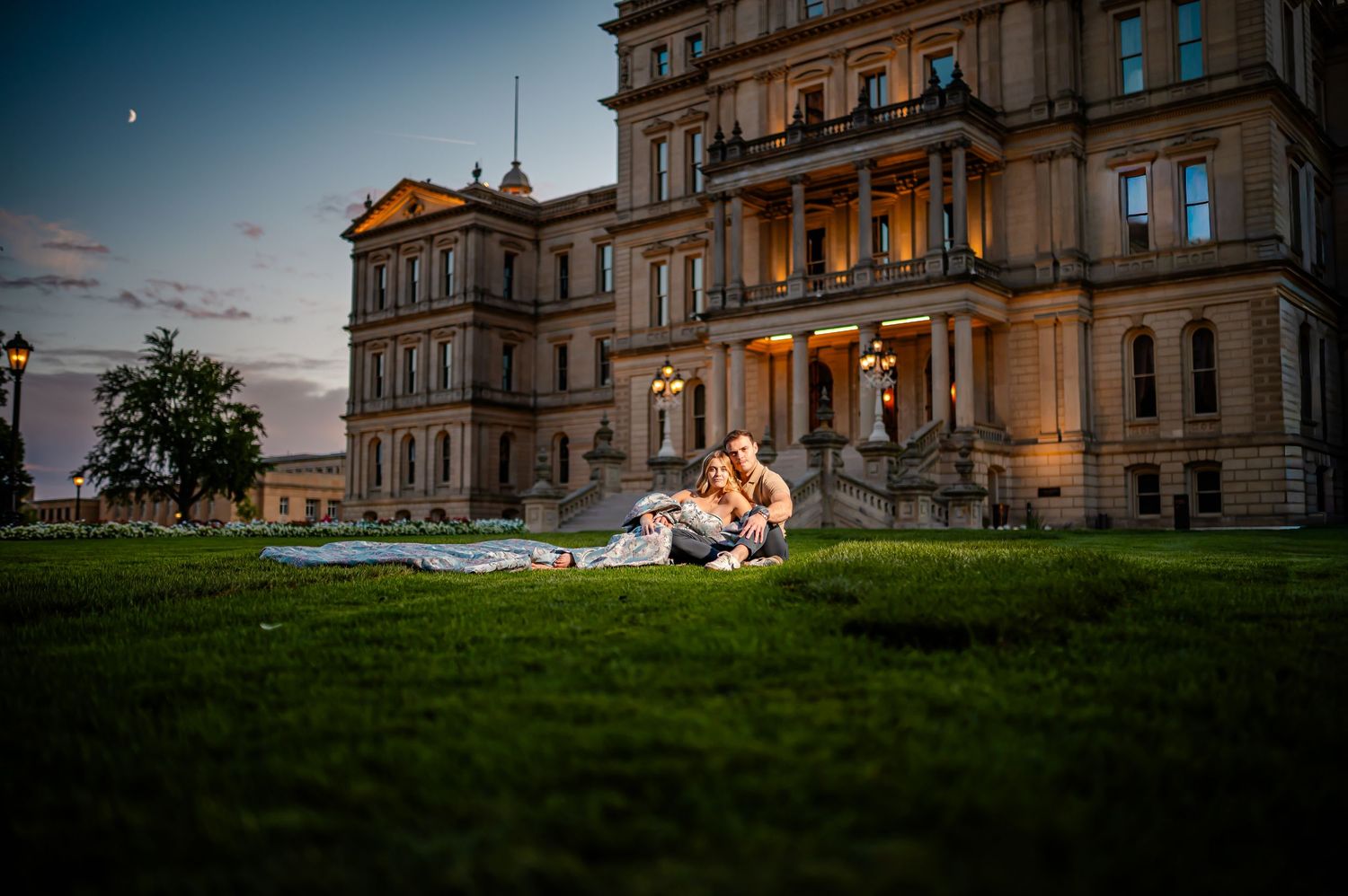 Two people enjoy a sunny evening on the lawn outside a grand stone mansion during sunset.