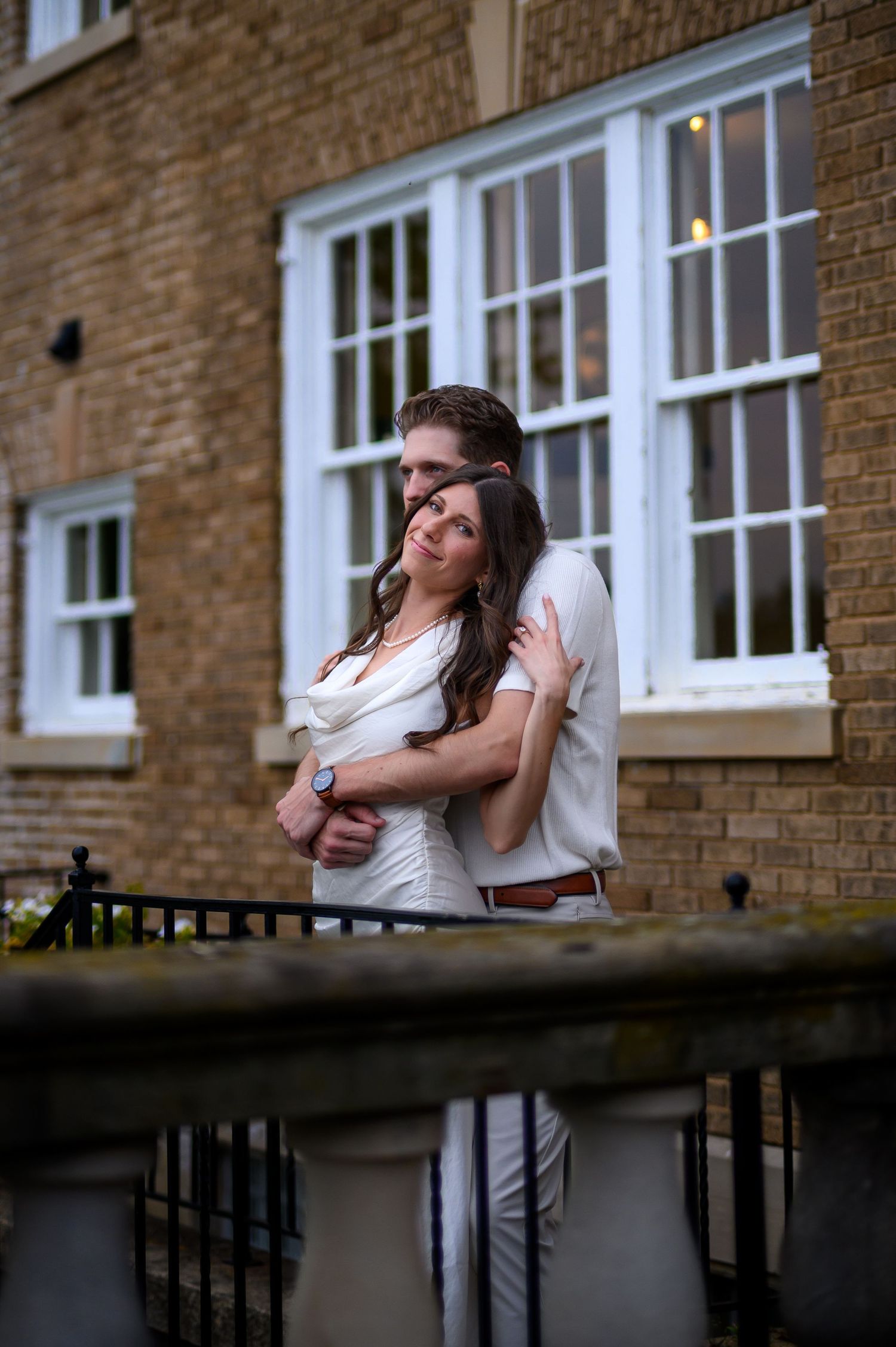 A romantic outdoor portrait of a couple in white outfits embracing against a brick building with tall windows.