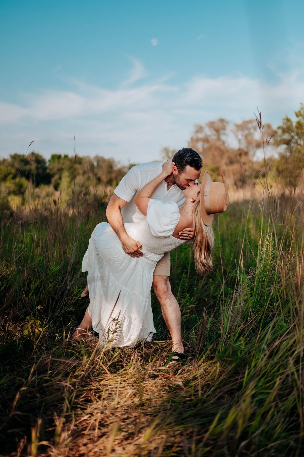 Romantic couple dips in a scenic outdoor engagement shoot amidst tall grass and blue skies at golden hour.