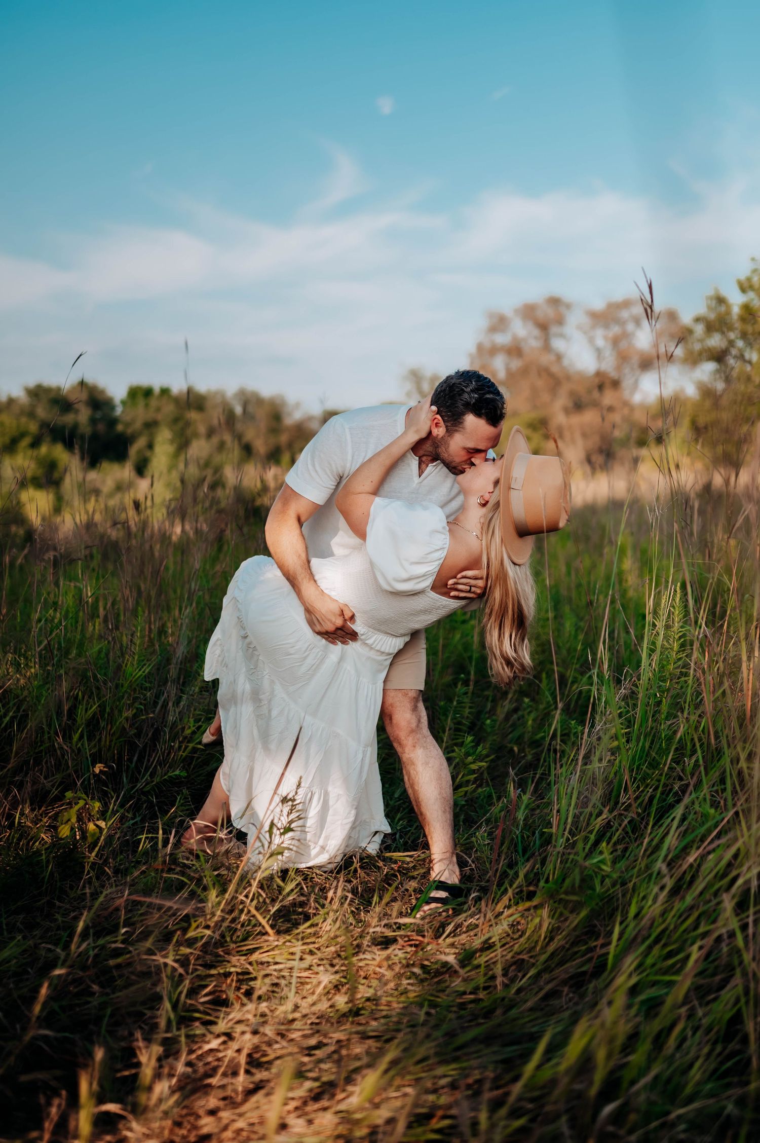 Romantic couple dips in a scenic outdoor engagement shoot amidst tall grass and blue skies at golden hour.