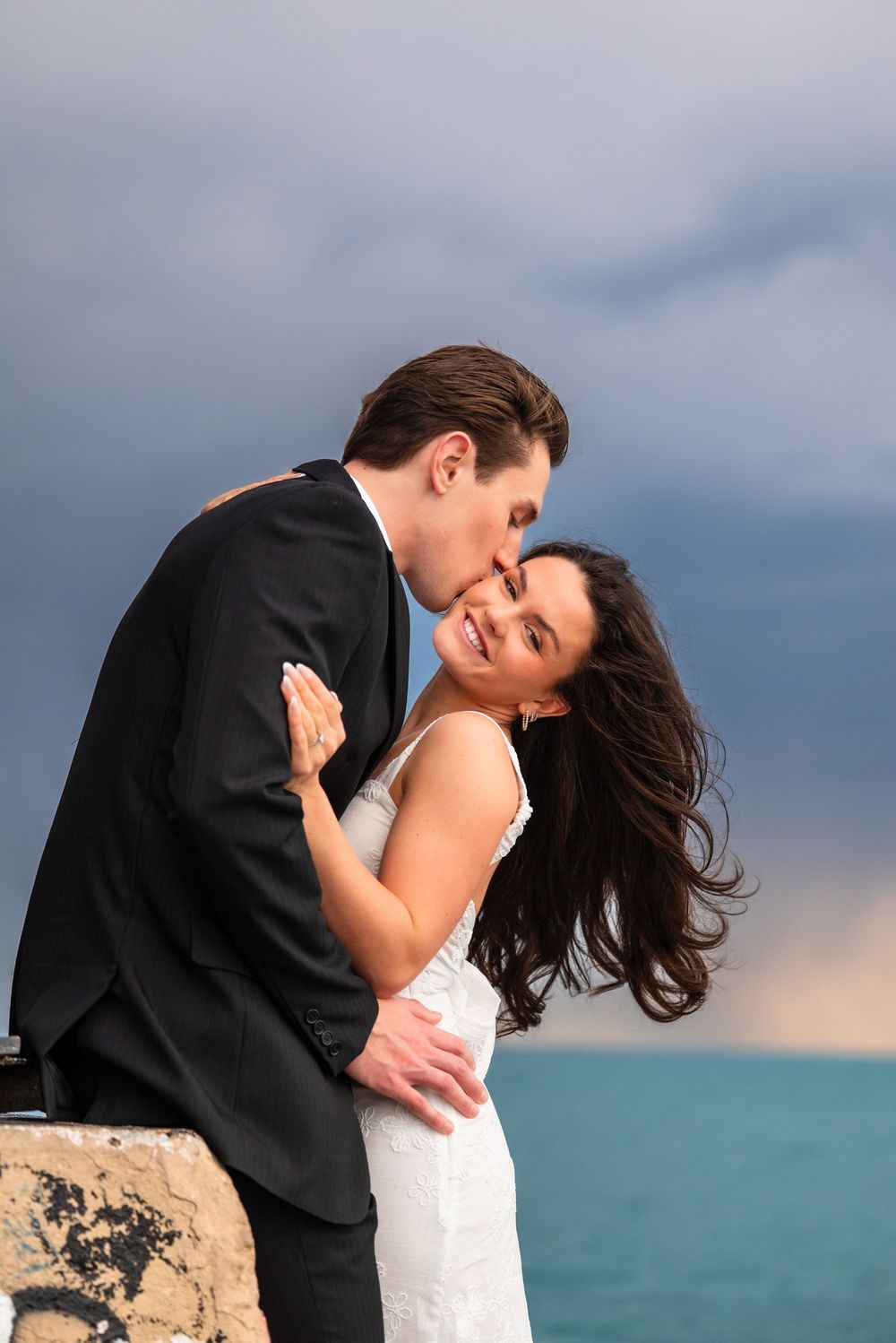 A groom in a black suit kisses his smiling bride in a white dress by the ocean under a dramatic cloudy sky.