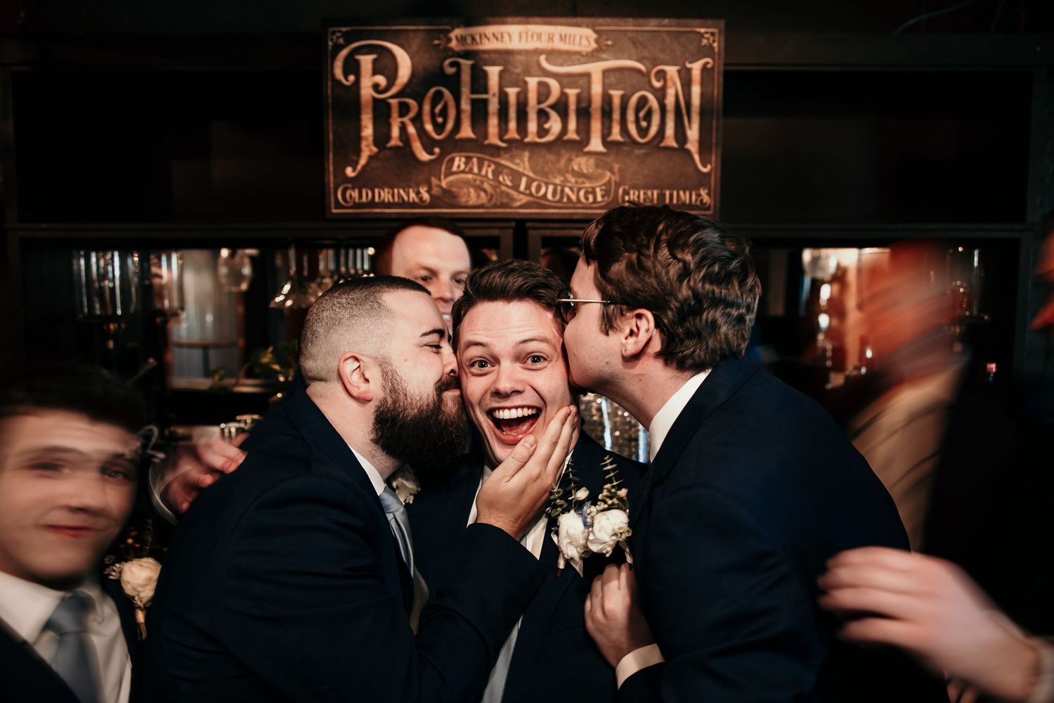 Groom gets kissed on cheeks by groomsmen in front of Prohibition Bar lounge sign.