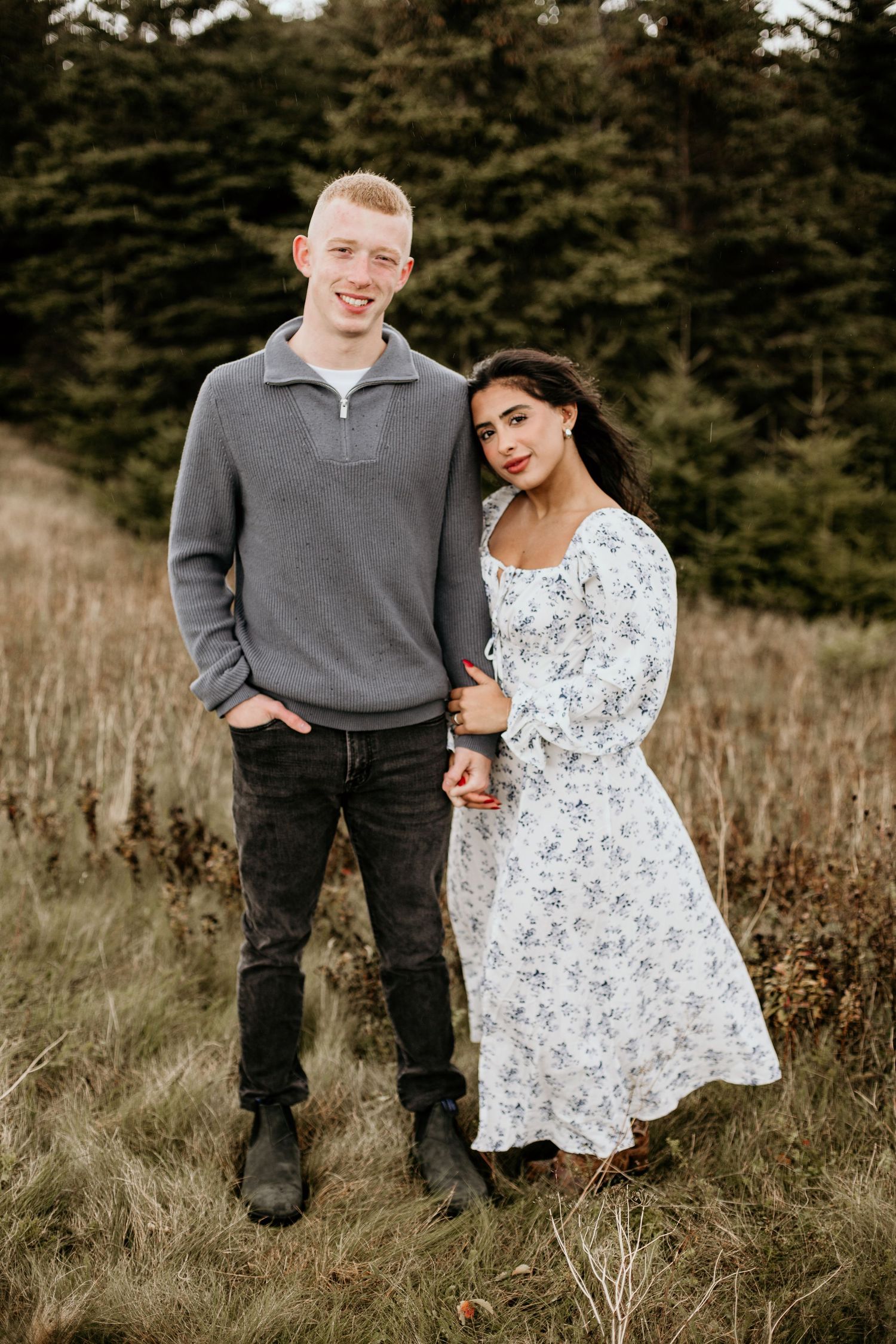 woman holding man's hand while resting her head on his shoulder, looking at the camera