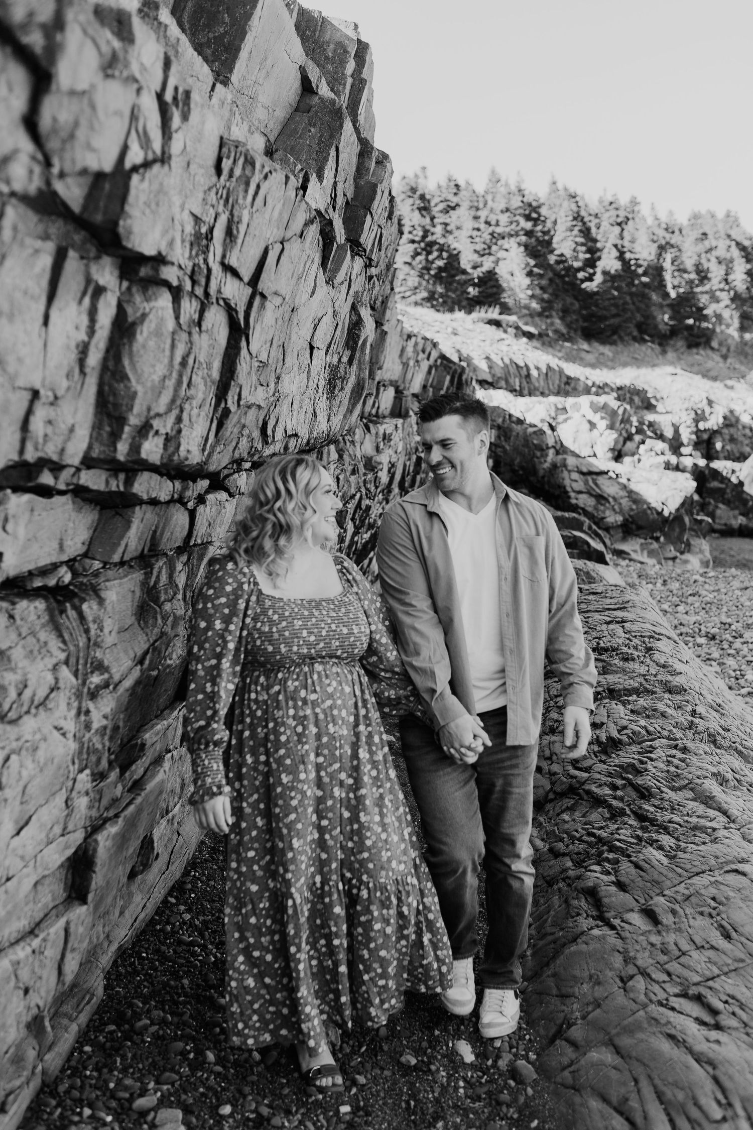 couple holding hands walking near a wall of rocks on a beach, looking and smiling at each other