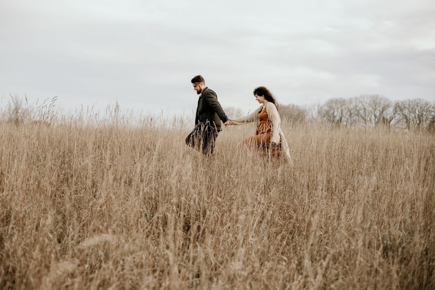 married couple walking through field of long grass, holding hands, man leading woman