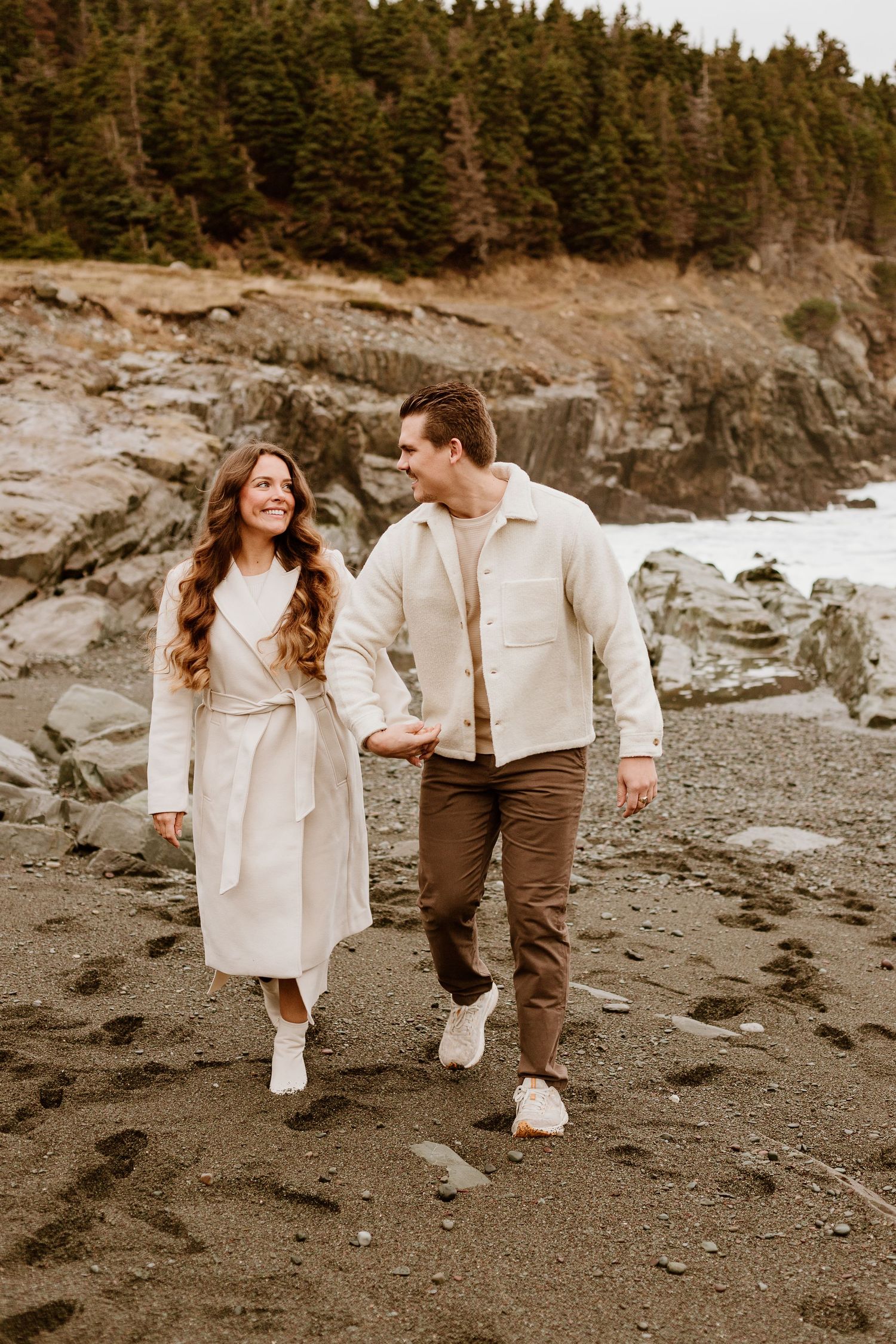 newly engaged couple holding hands walking on the beach smiling at each other