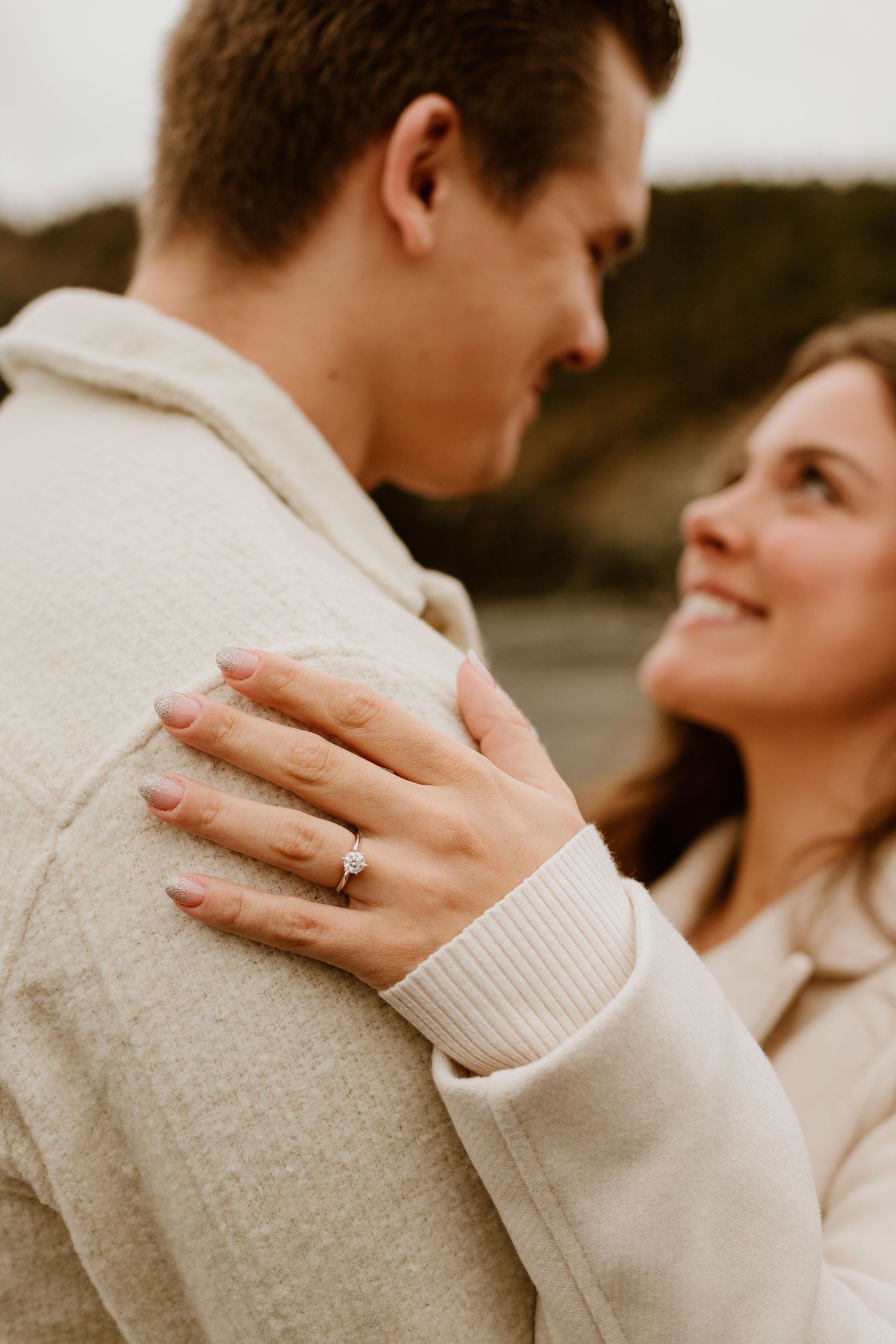 close up shot of a woman's engagement ring with the engaged couple looking at each other in the background