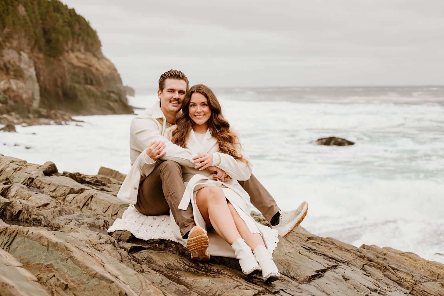 engaged couple cuddling on a rock by the beach