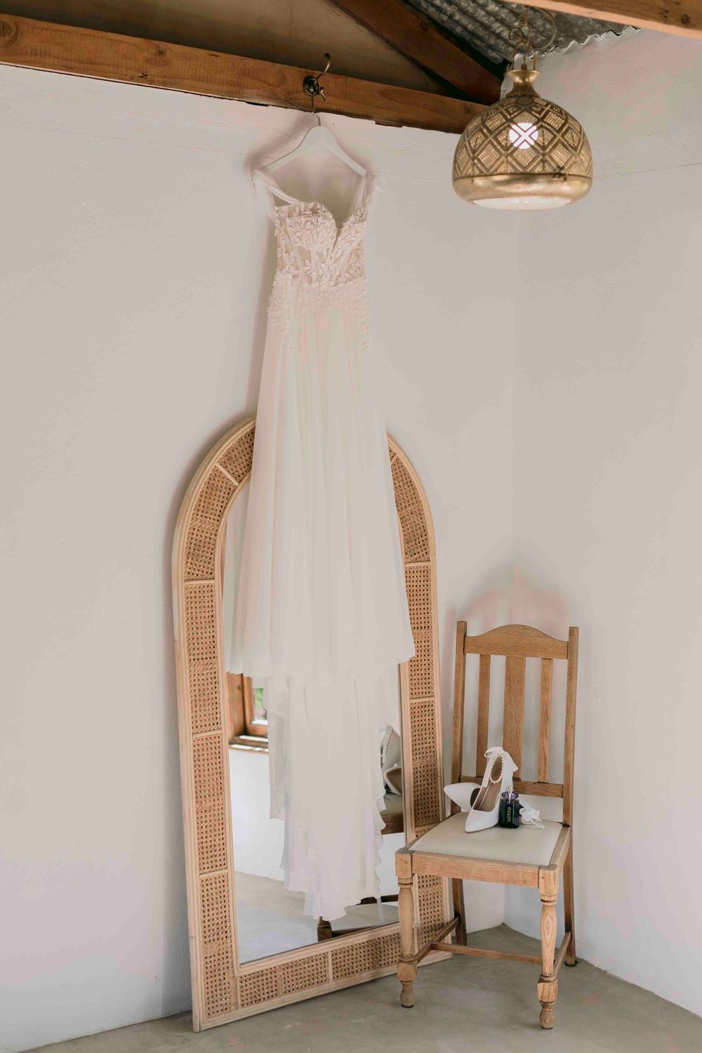 Elegant white lace wedding dress hanging on a rattan arch mirror beside a wooden chair with a Moroccan pendant light.