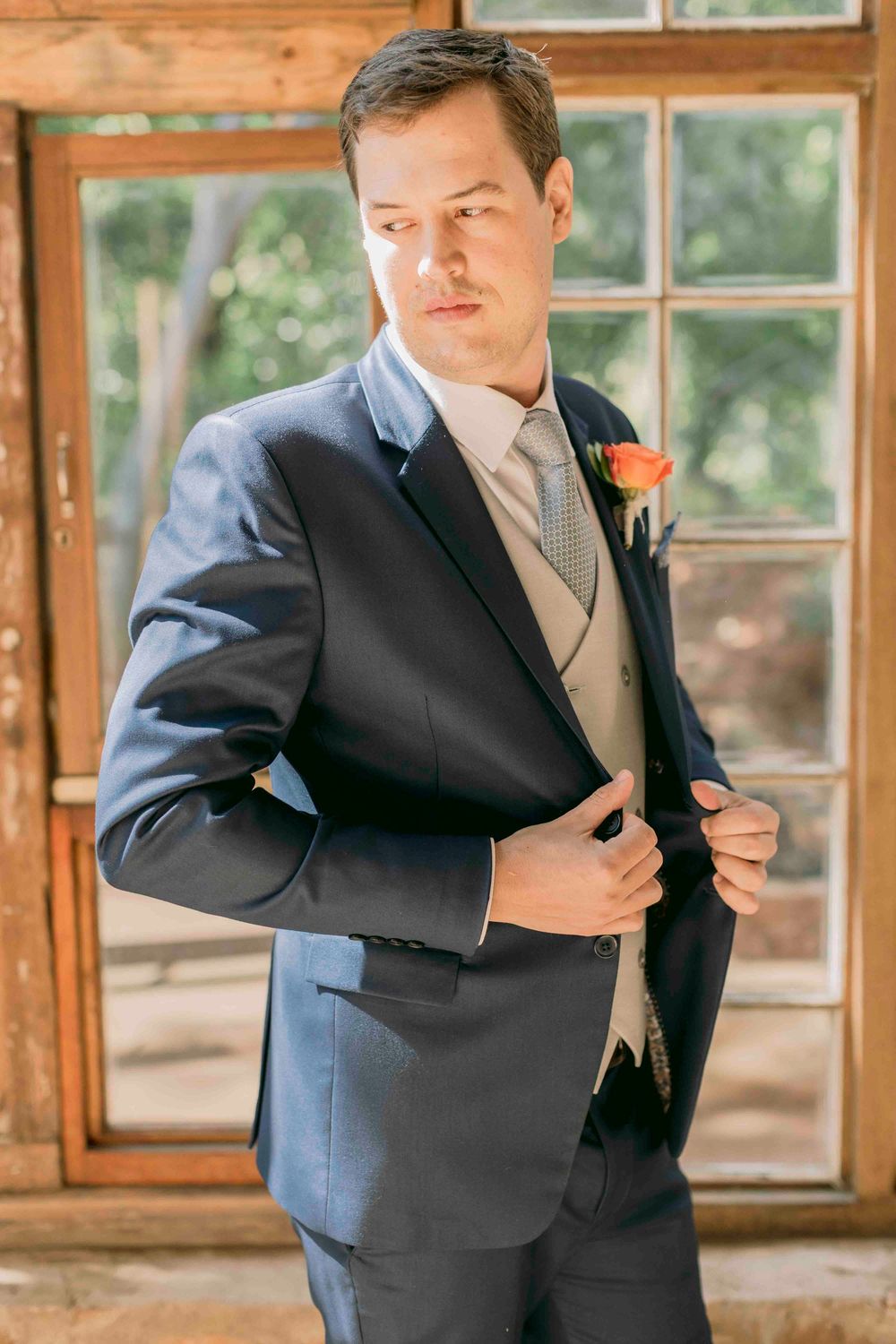 Dapper groom in navy suit with beige vest and orange boutonniere adjusting his jacket indoors.